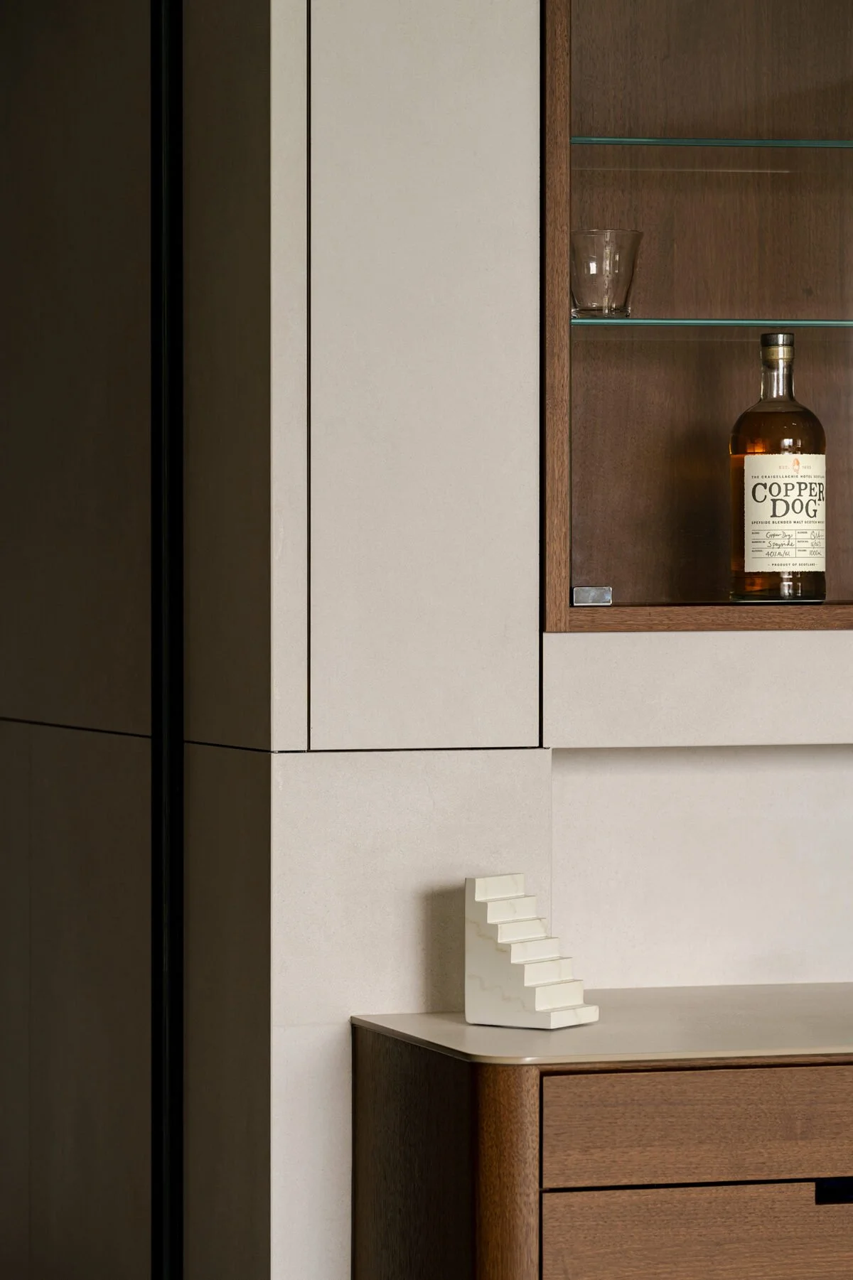 A corner of a modern room with beige and wooden cabinets, a glass shelf with a glass and a whiskey bottle labeled 'Copper Dog', a small beige stepped sculpture, and an electrical outlet on the wall.