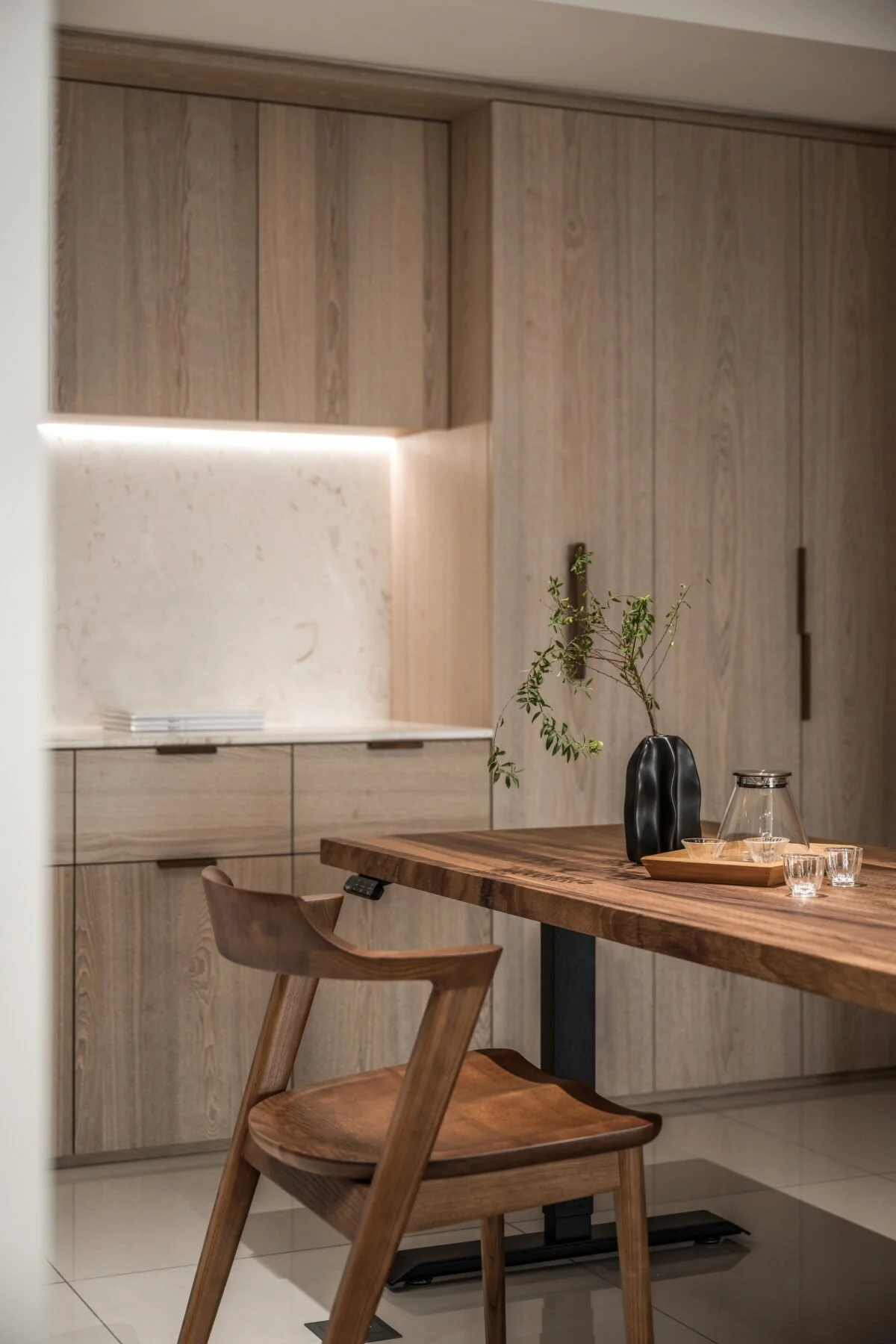 A modern kitchen with a wooden dining table, a black vase with green branches, and glassware. There are wooden cabinets and a beige backsplash. A wooden chair is in the foreground.