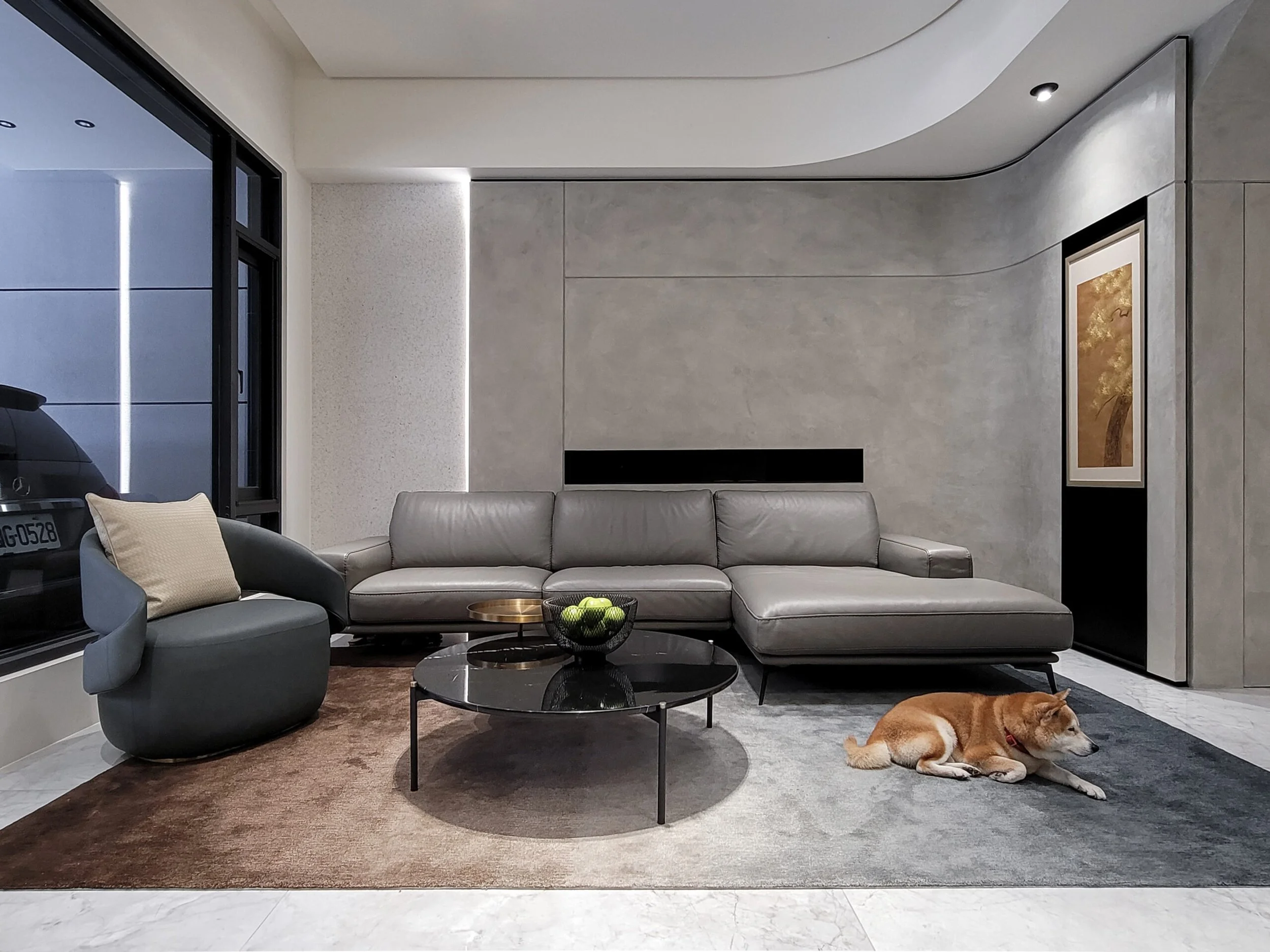 Modern living room with gray leather sofa, black coffee table with green apples in a basket, a black armchair with a beige pillow, a cat rug, and a brown and white dog lying on the floor. There is a large framed artwork on the wall, a window with a black frame, and built-in lighting.