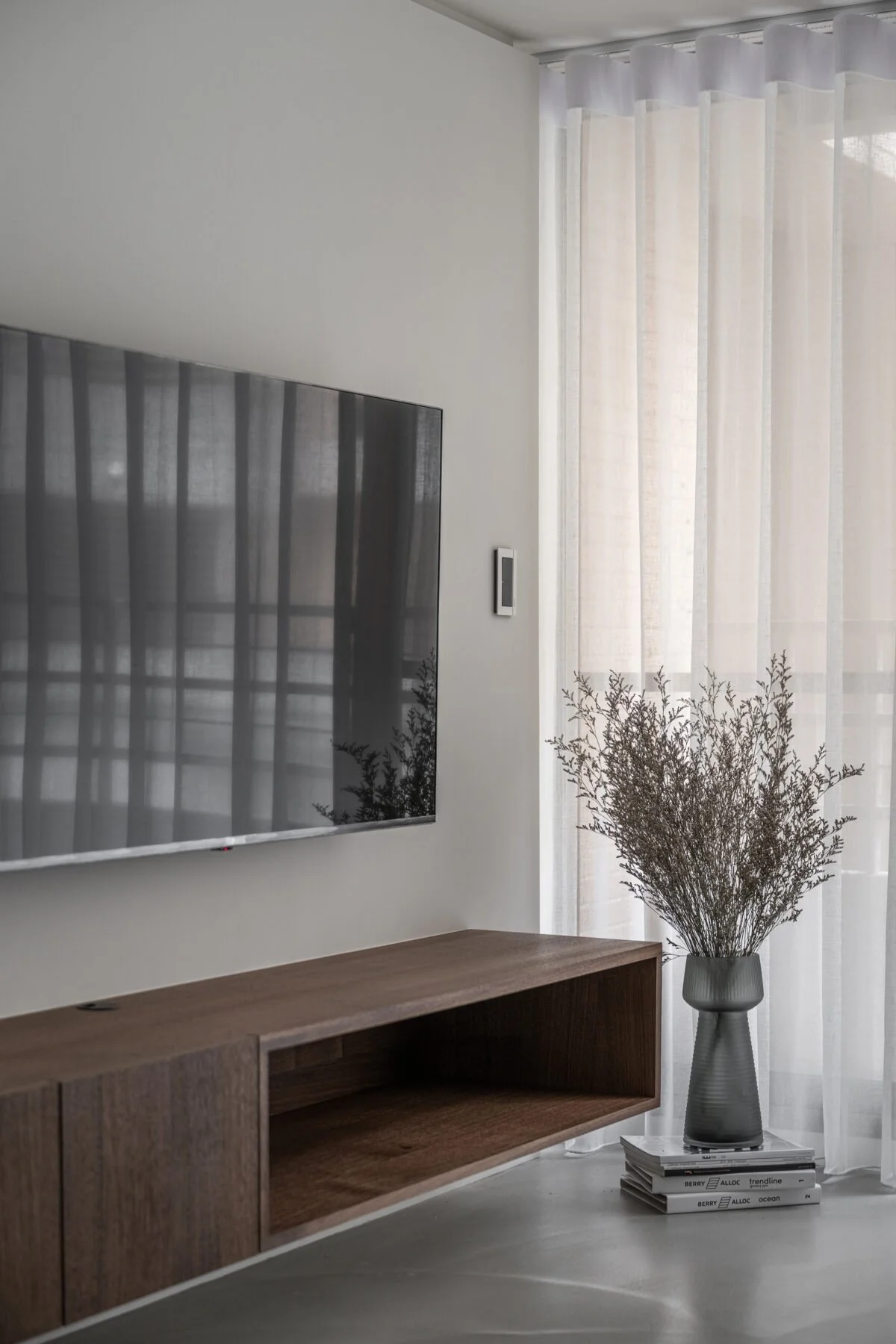 A modern living room with a wall-mounted television, sheer curtains, a wooden console, a tall vase with dried branches, and books stacked on the floor.