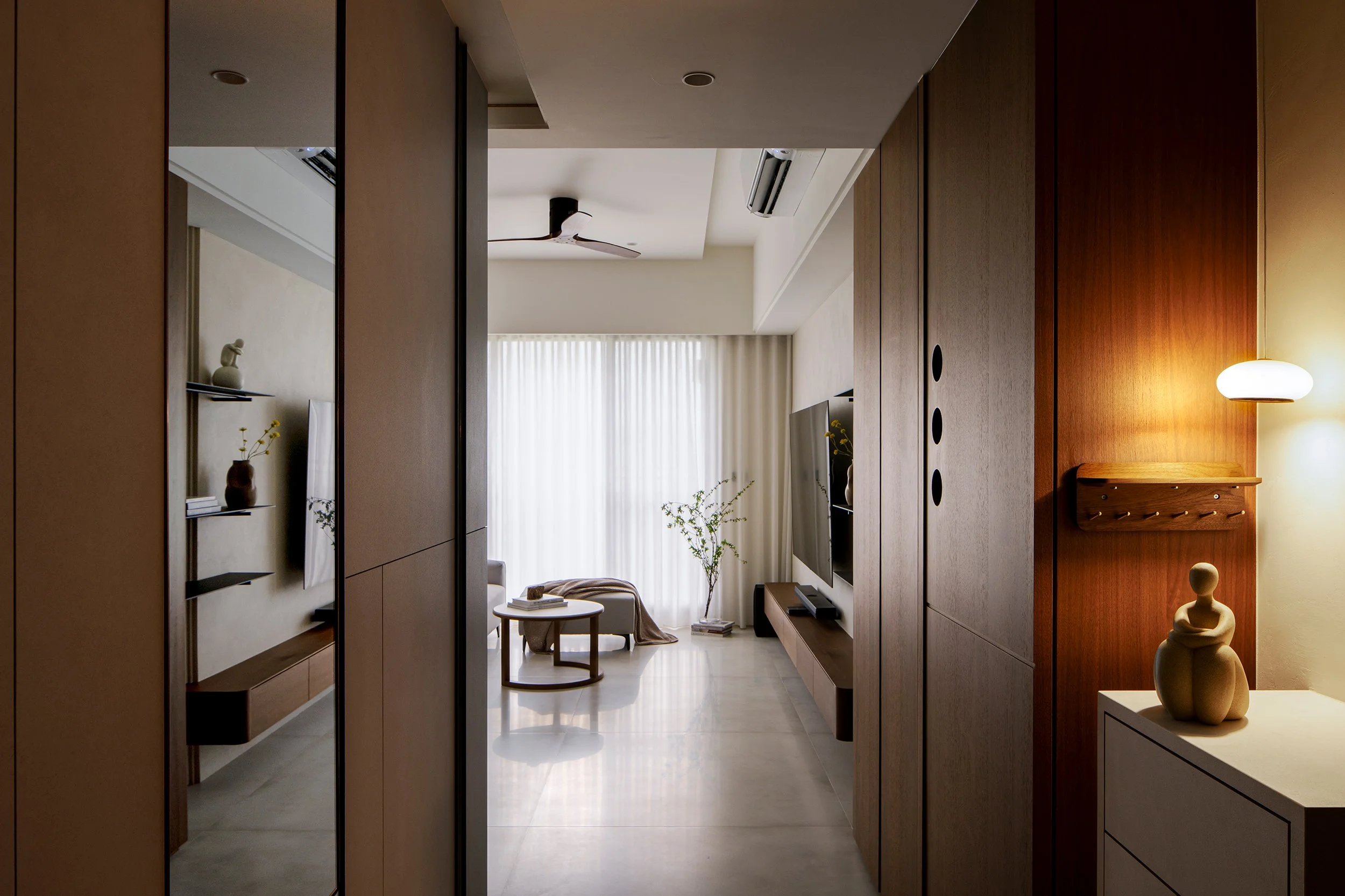 Living room with wooden walls, a wall-mounted TV, a white curtain, a ceiling fan, and a coffee table with books and decor, viewed through a doorway with mirrored panels.