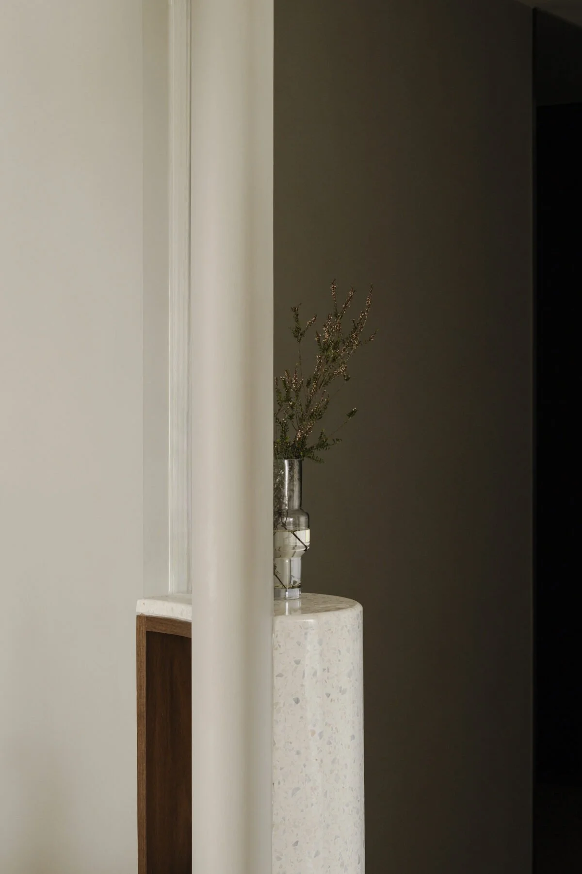 A minimalist interior design scene showing a white marble round side table with a clear glass vase holding dried lavender plants, positioned next to a wall, with a partial view of a wooden shelf.
