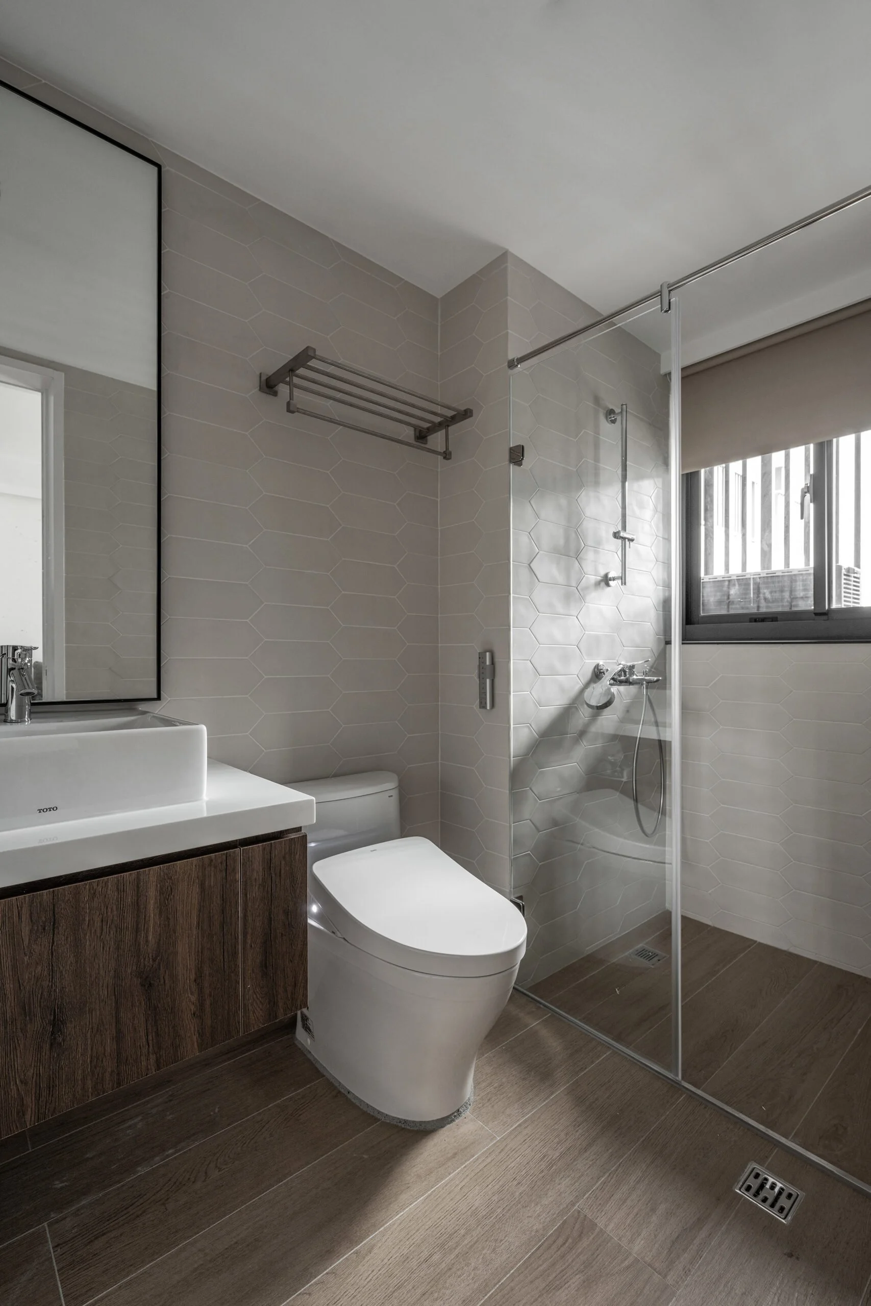 Modern bathroom with beige hexagonal tile walls, a white toilet, a wooden vanity with a white sink and mirror, a glass shower enclosure with a rainfall showerhead, a window with blinds, wood-look floor tiles, and a floor drain.