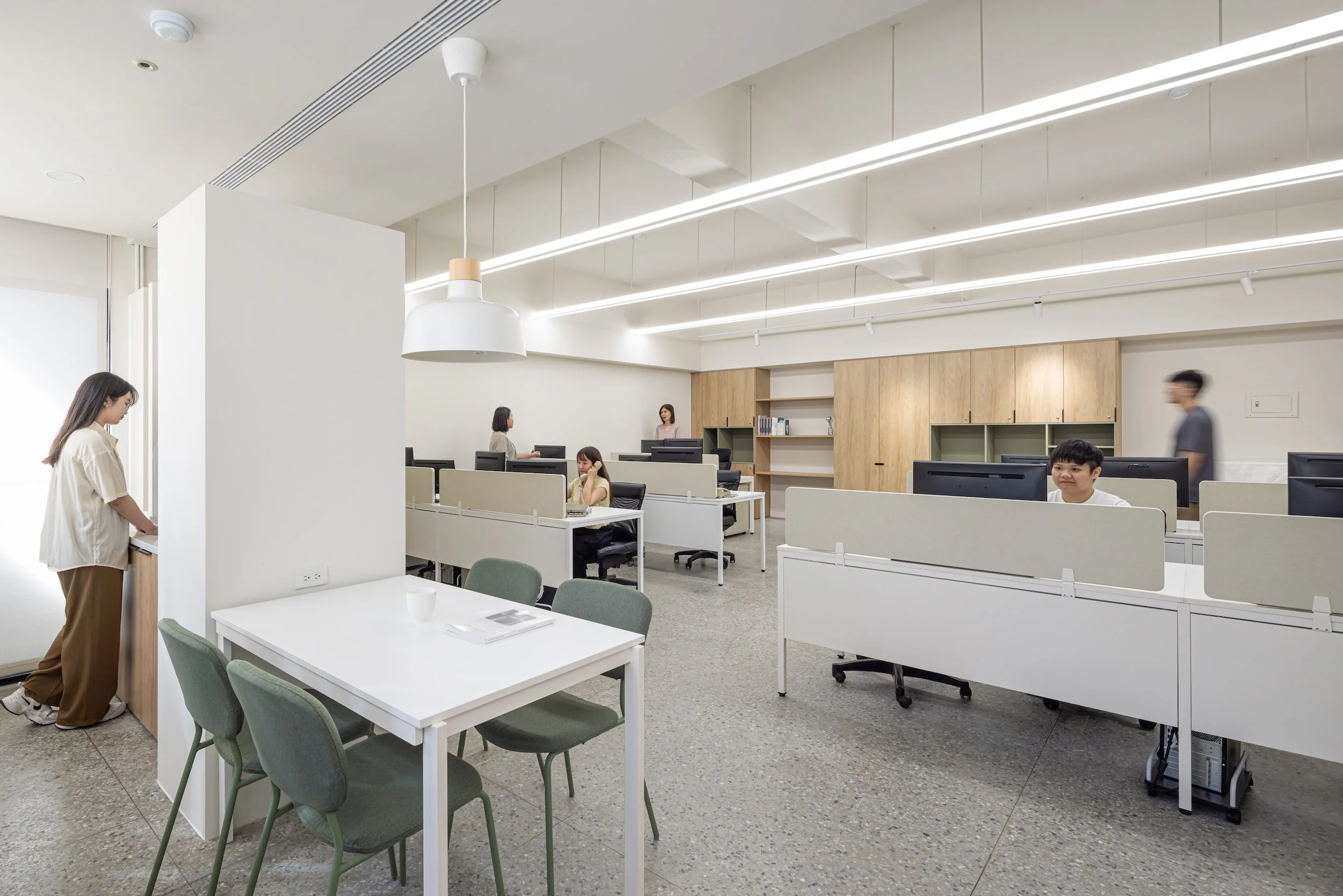 Modern office space with white desks and chairs, several people working at computers, and a wooden wall with shelves, bright lighting, and a few people engaged in different tasks.