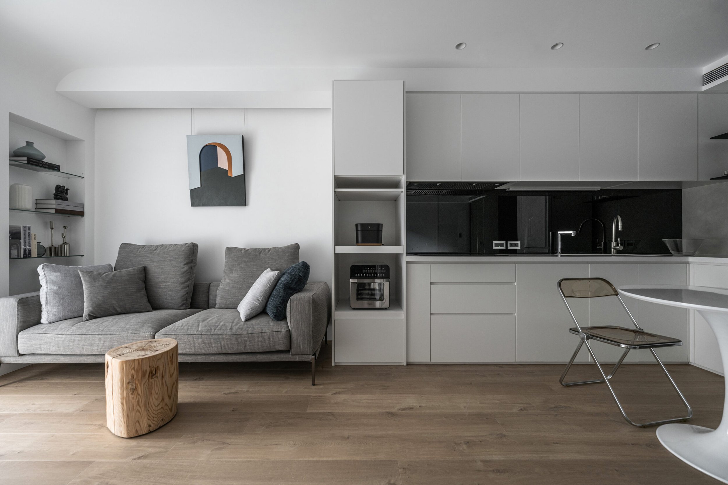 Modern living room and kitchen with gray sofa, wood coffee table, white cabinets, black backsplash, under-cabinet lighting, decorative shelves, and wood flooring.