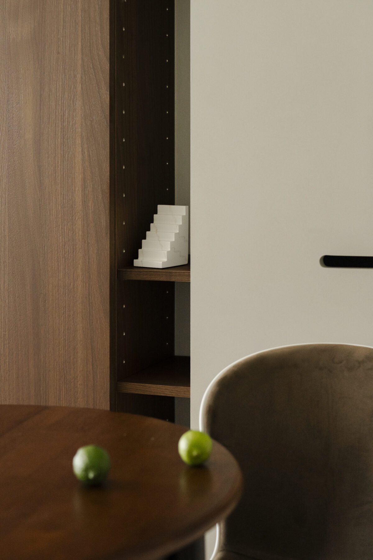 Part of a living room or dining area showing a wooden table with two green grapes on it, a gray and brown chair, a wooden bookshelf with a white staircase figure on one of its shelves, and a white wall.