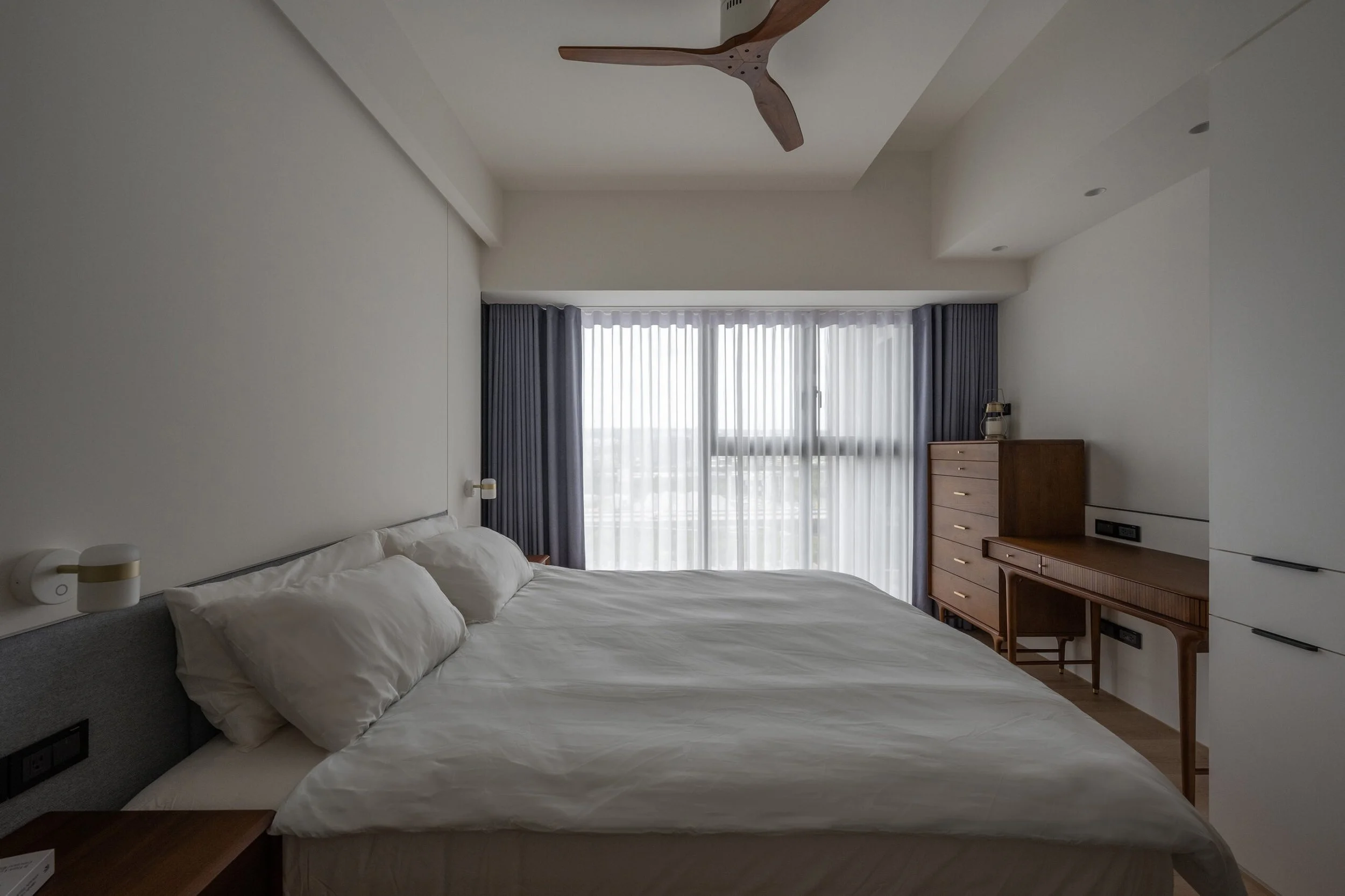 A bedroom with a large bed, white bedding, and pillows, next to a window with sheer curtains and dark drapes, a wooden dresser, and a ceiling fan.