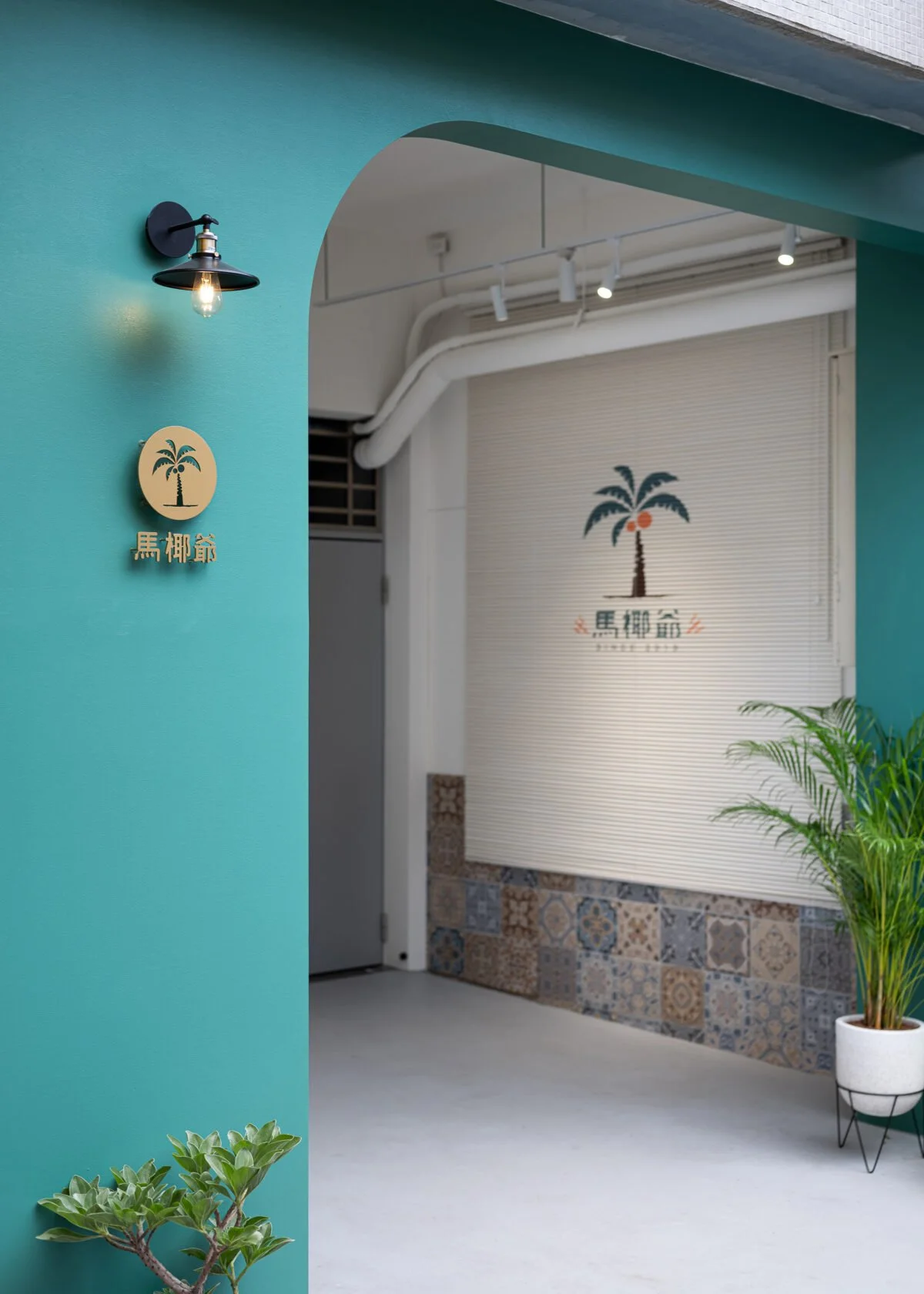 Entrance with teal wall, black wall sconce light, gold palm tree sign with Chinese characters, decorated roller shutter with palm tree logo, potted plant, and small green shrub.