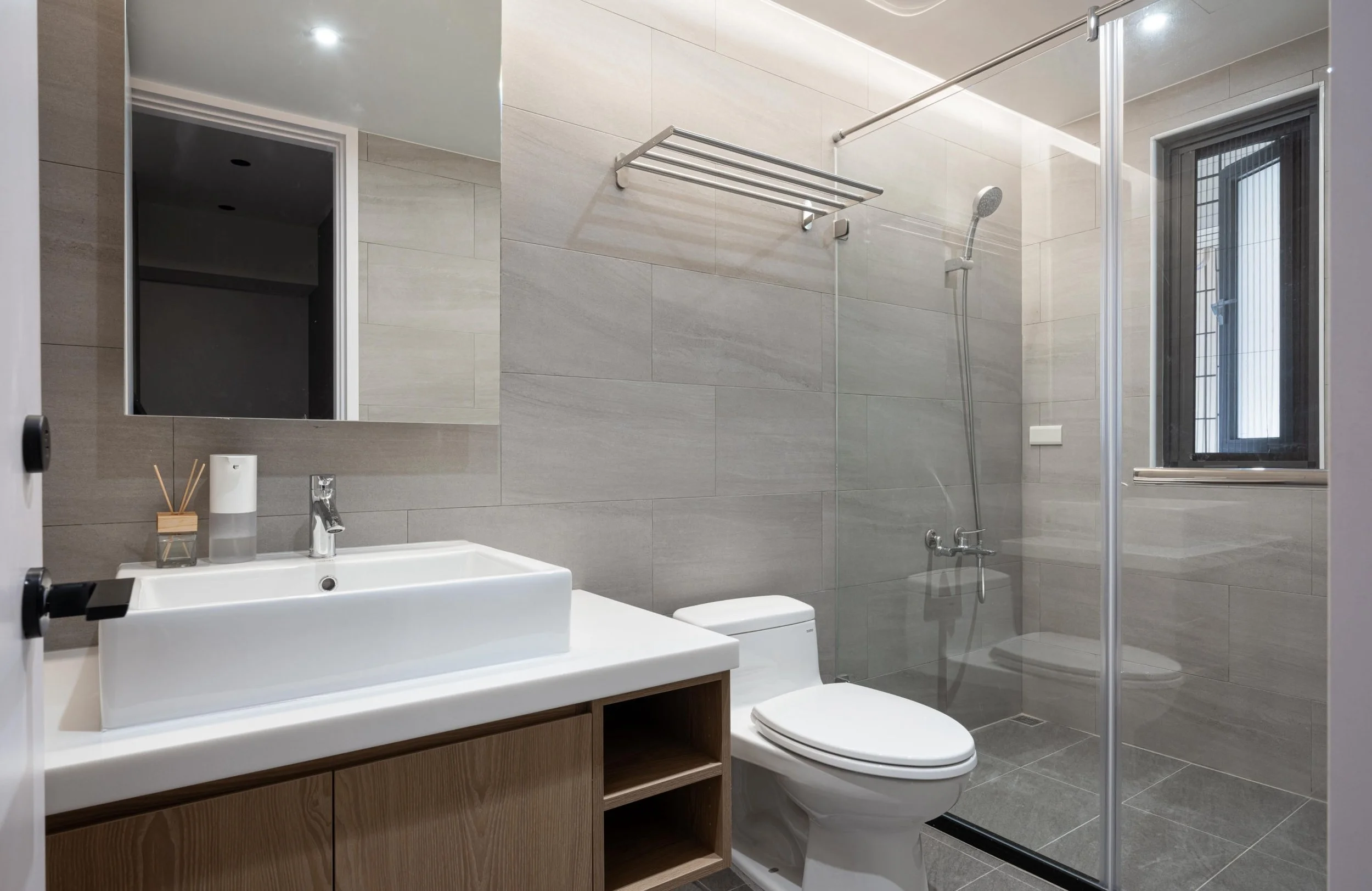 Modern bathroom with gray tiled walls, a white sink with a wooden vanity, a mirror, a toilet, a shower area with a glass door, a window, and towels hanging on a rail.