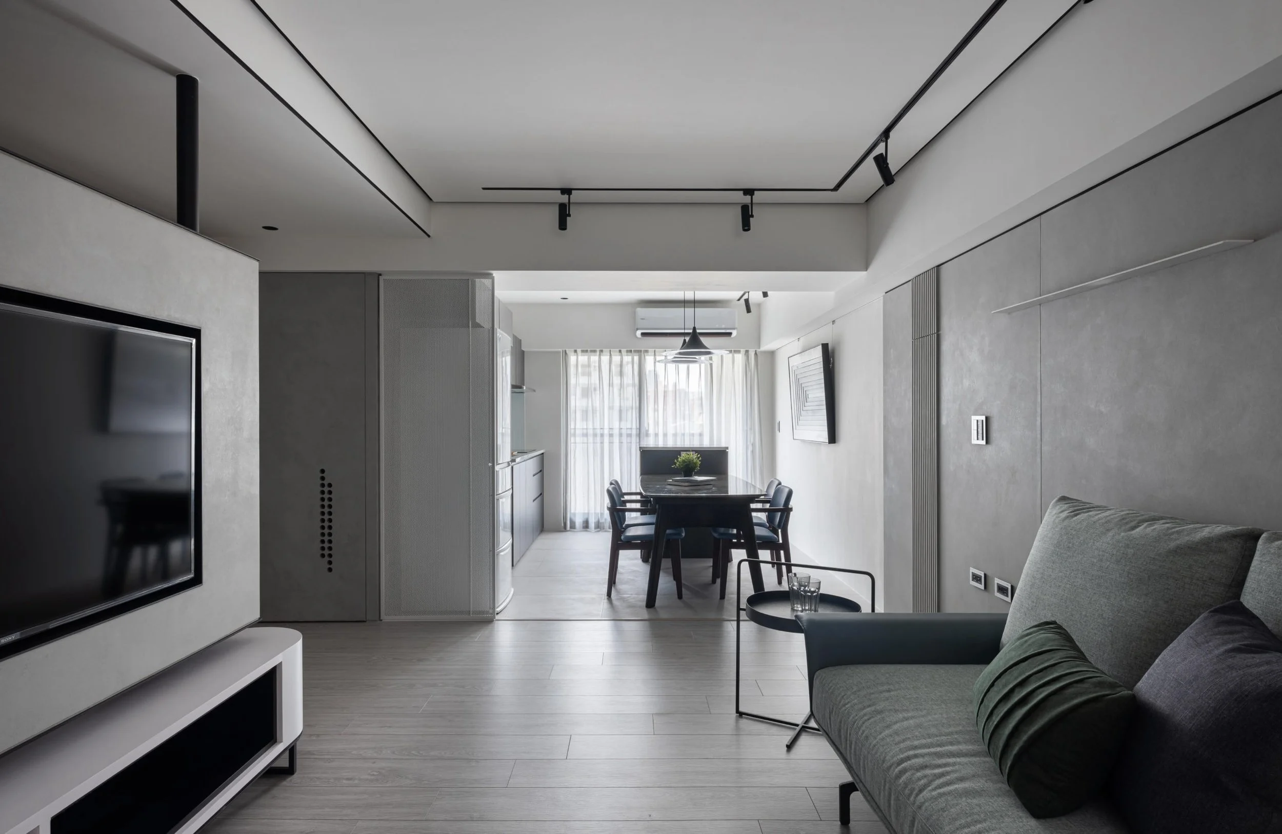 Modern living room with gray sofa, black side table, wall-mounted TV, and dining area with black table, chairs, and a large window with white curtains in the background.