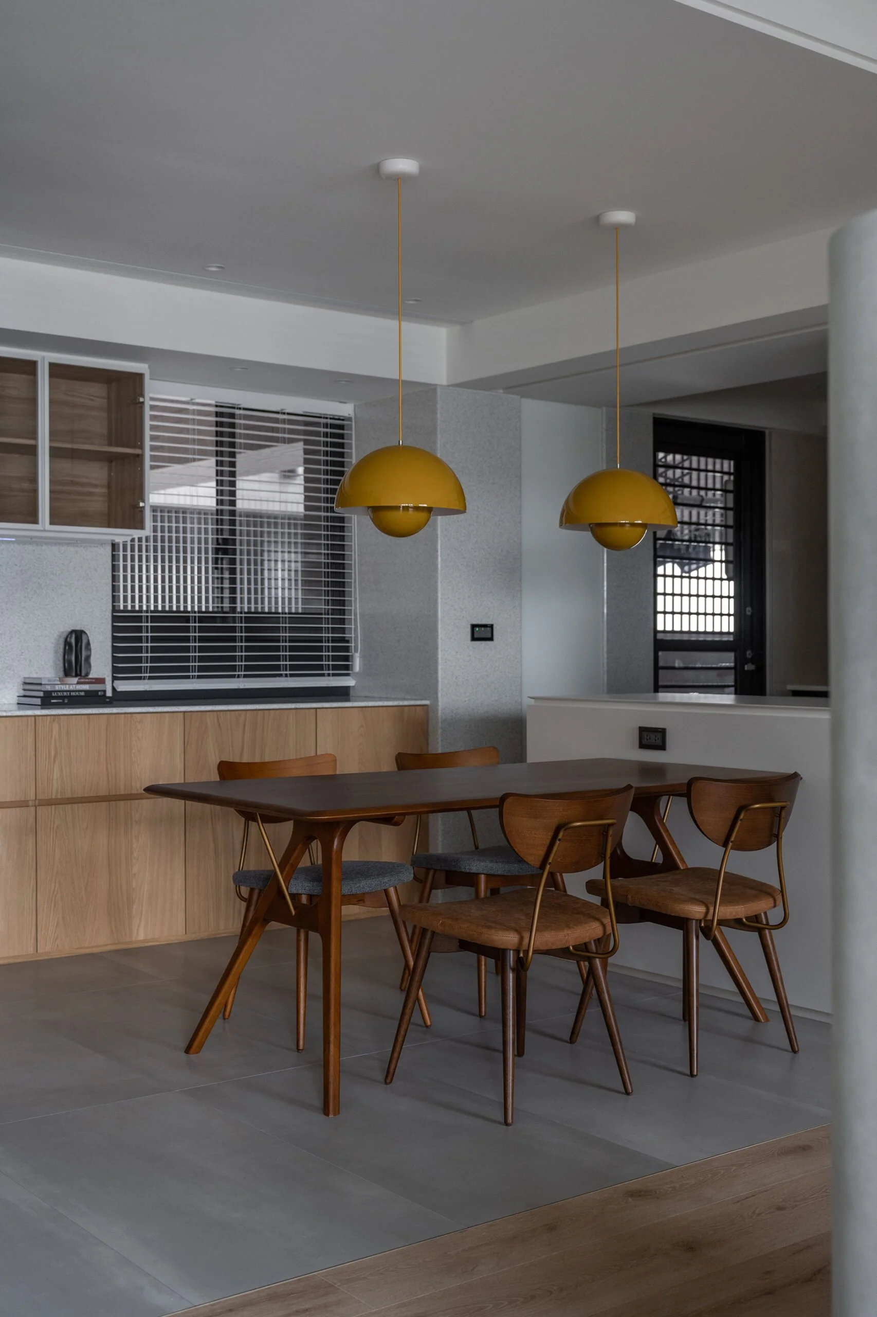 Modern dining area with a dark wood table, five wooden chairs with upholstered seats, two yellow pendant lights hanging from the ceiling, a cabinet with wooden doors, a large window with white blinds, and a stone wall section.