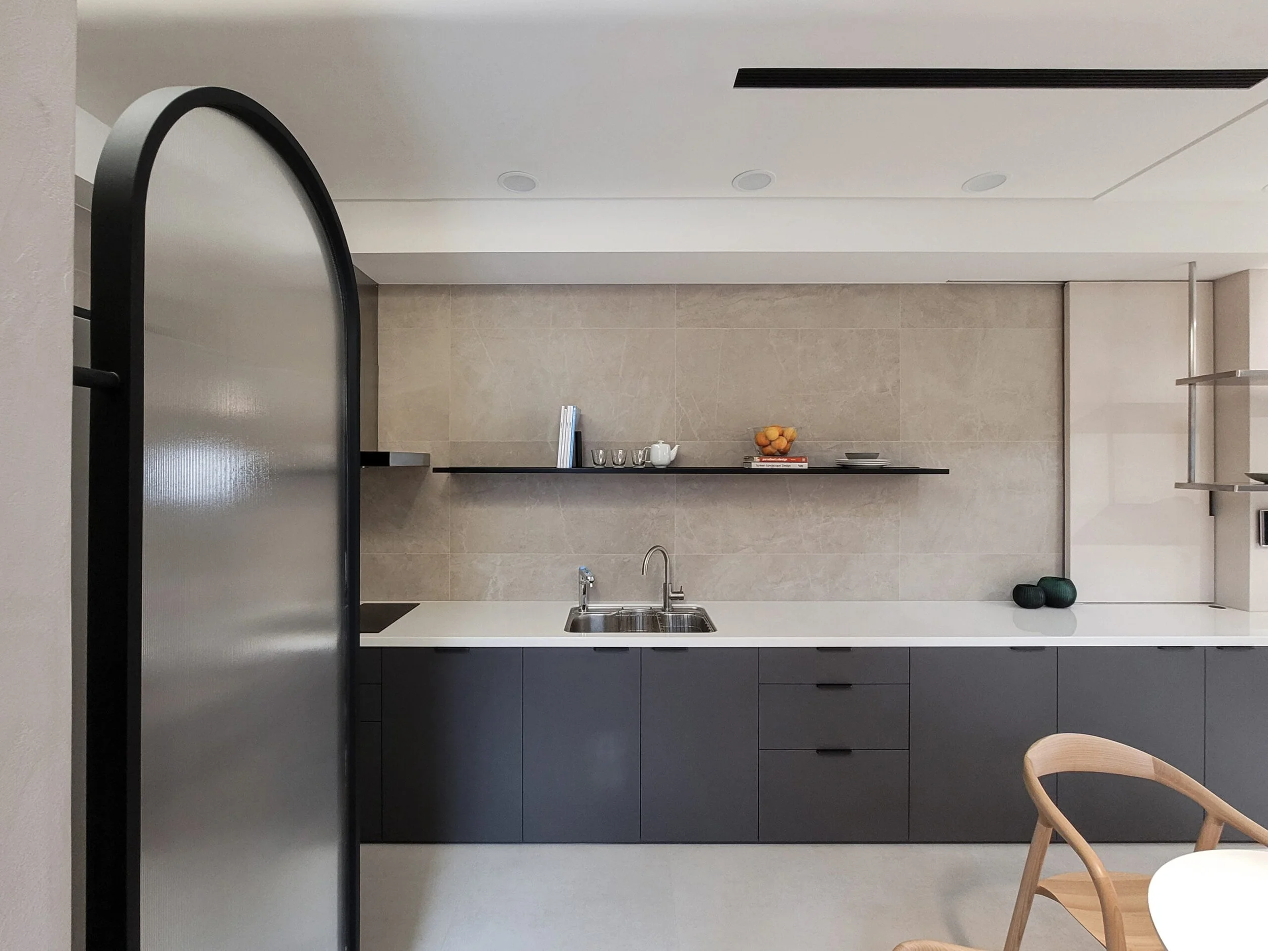Modern kitchen with beige tiled backsplash, black lower cabinets, and minimal decor including a white pitcher, oranges, and stacked plates. A wooden chair is partially visible in the foreground.