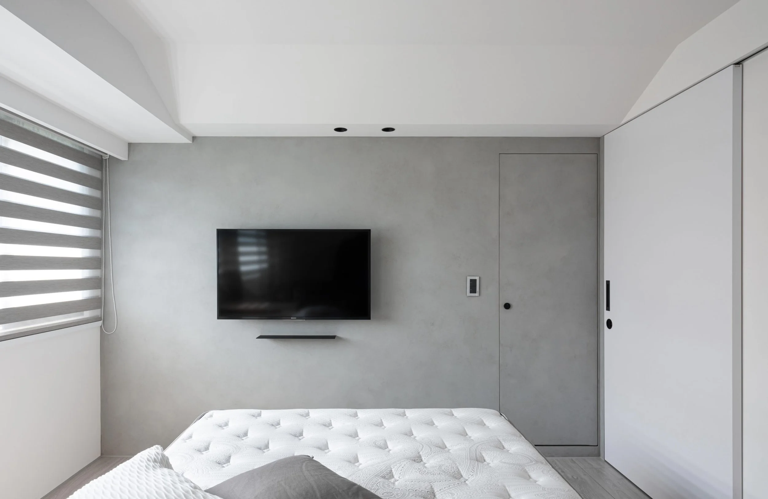 Modern minimalist bedroom with a wall-mounted TV, gray wall, window with horizontal blinds, a mattress with white quilted bedding, and a door, all in a neutral color scheme.