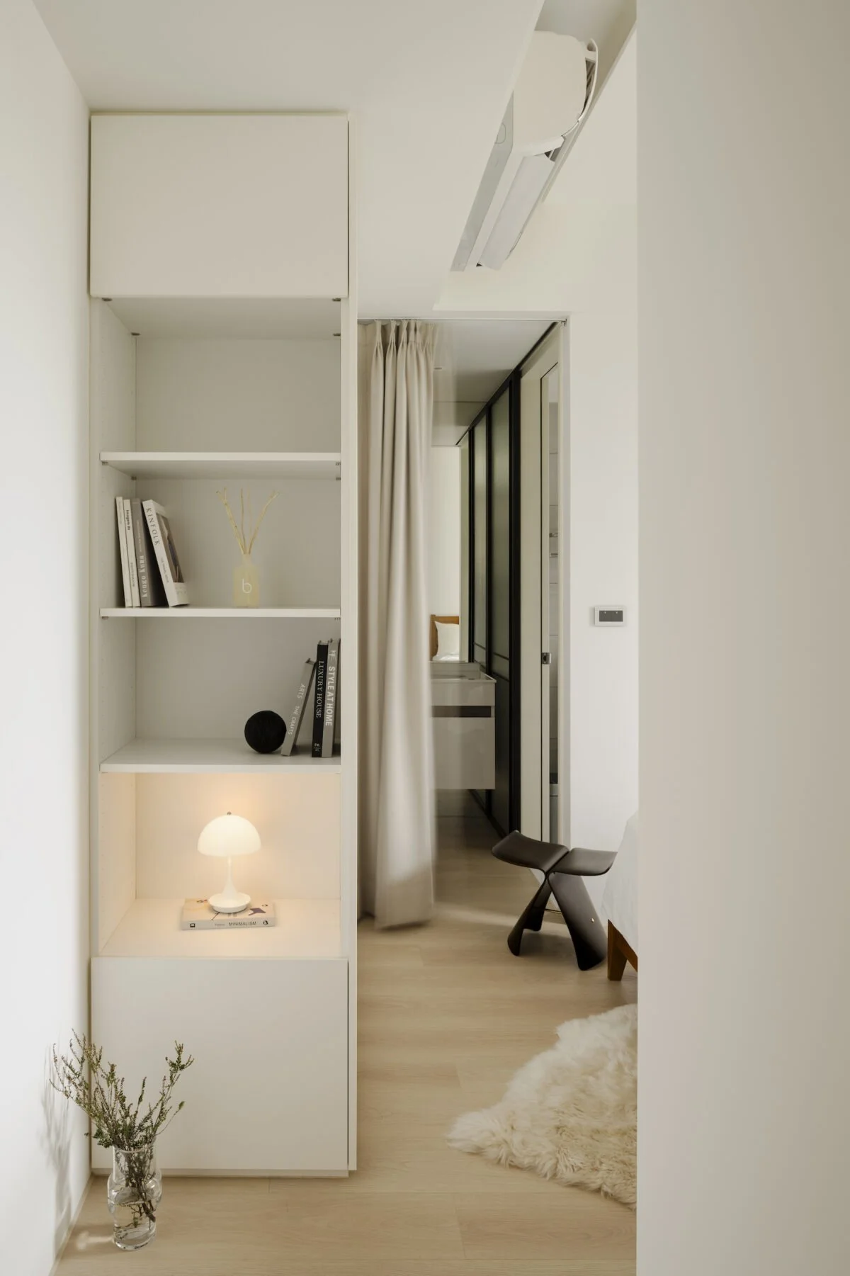 A minimalistic modern bedroom with a white bookshelf, a small table lamp, a vase with dried plants, a fluffy rug, and a black ergonomic chair near a sliding door to a bathroom or closet.