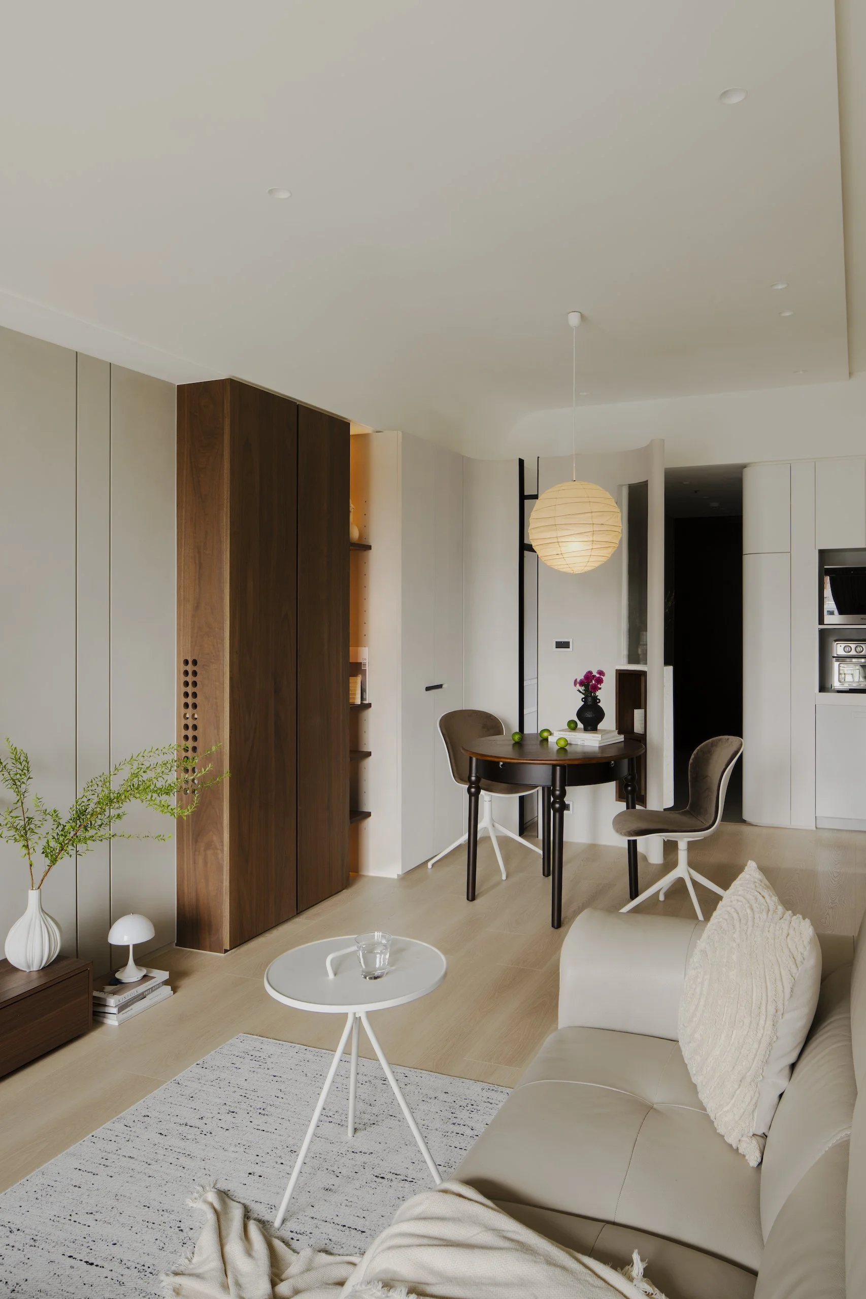 A modern living room with a beige sofa, a small round white side table with a glass of water, a white rug with black stripes, a dining area with a round wooden table, two beige chairs, contemporary white walls, a wooden cabinet, and decor including a white lamp, a black vase with pink flowers, and a potted plant.