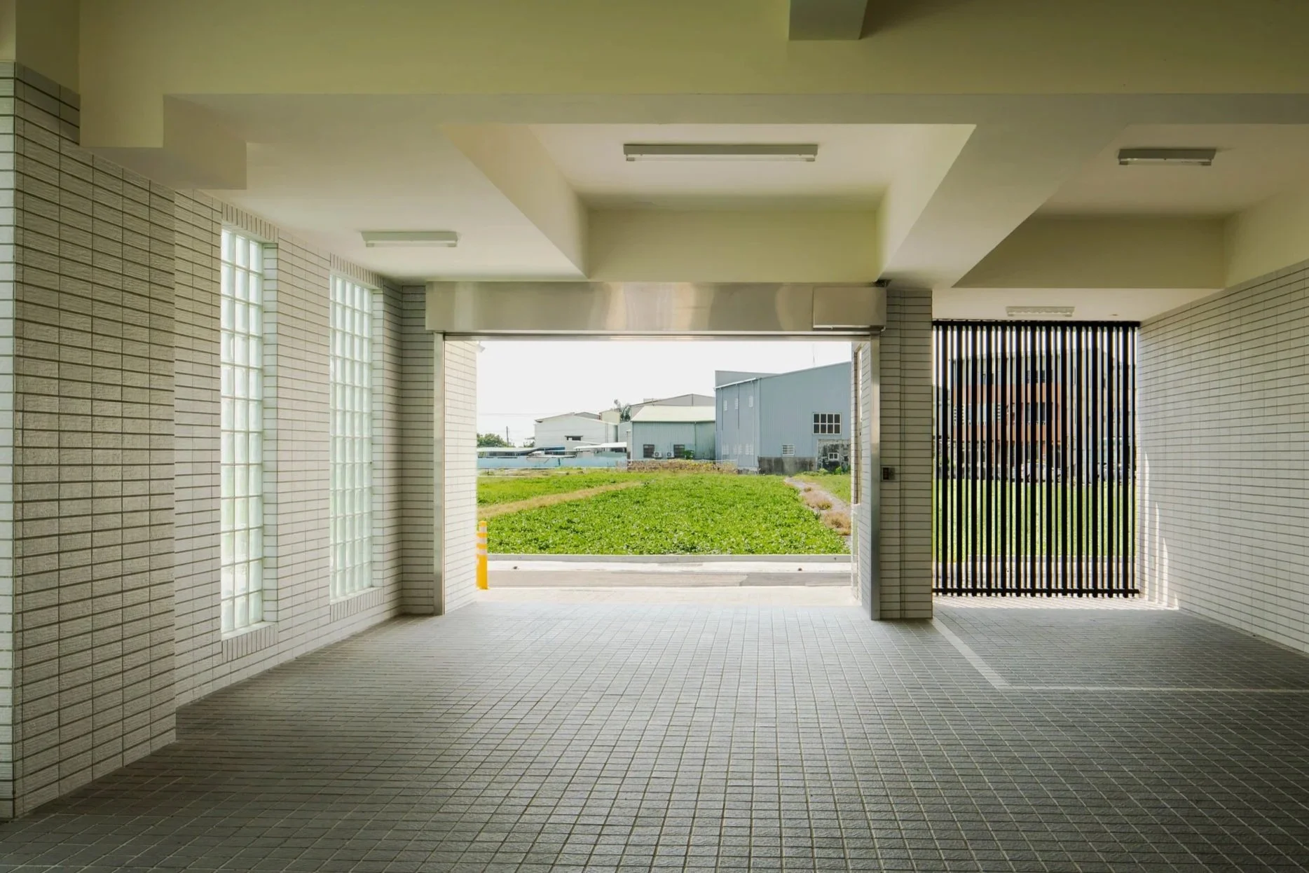 自遮蔽廊道下方往開闊處望去，視野穿過開放空間，遠處為草坪與工業廠房景致。