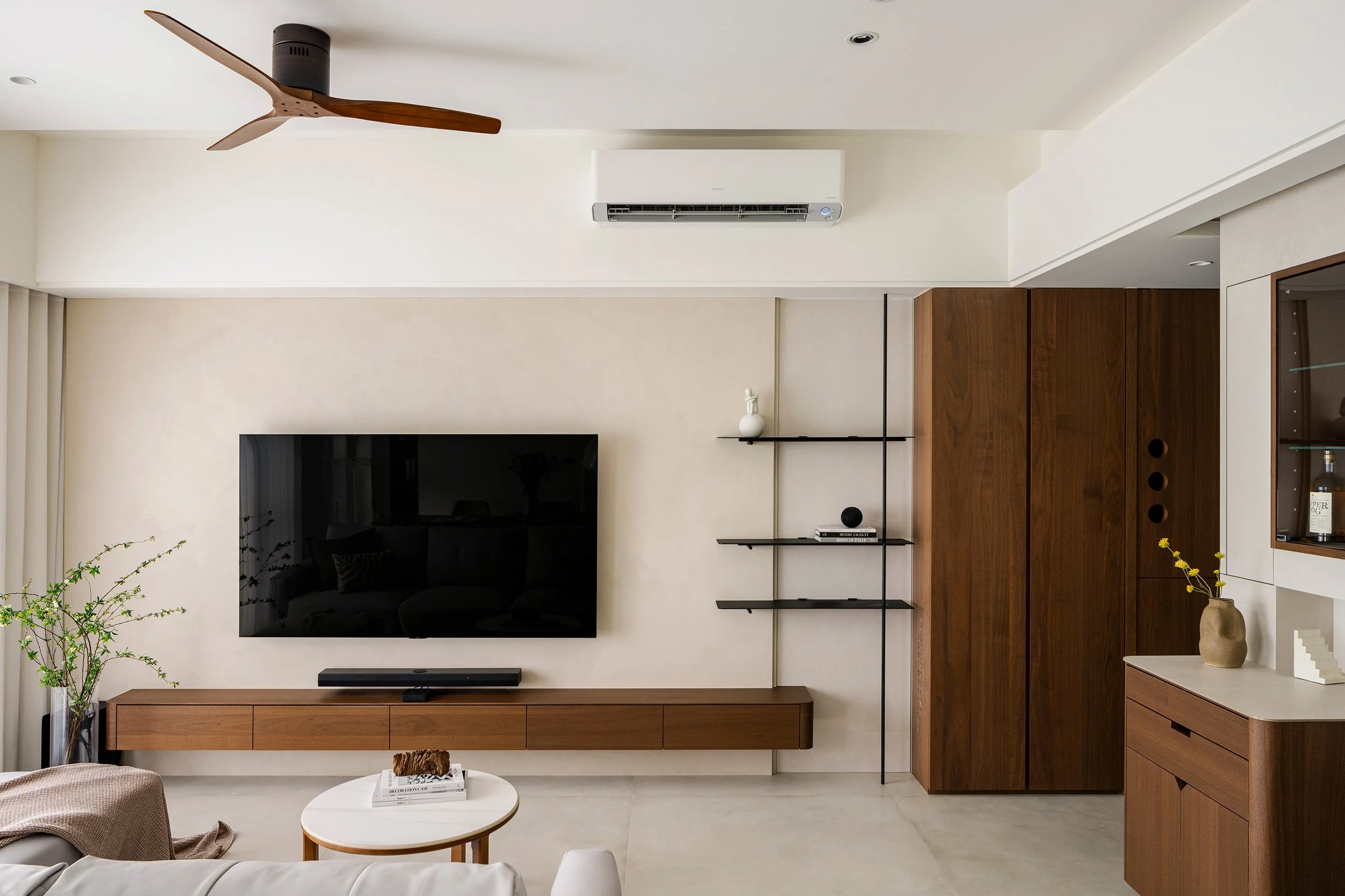Modern living room with a wall-mounted television, wooden entertainment unit, black shelves with decorative items, a ceiling fan, air conditioner, and a potted plant.