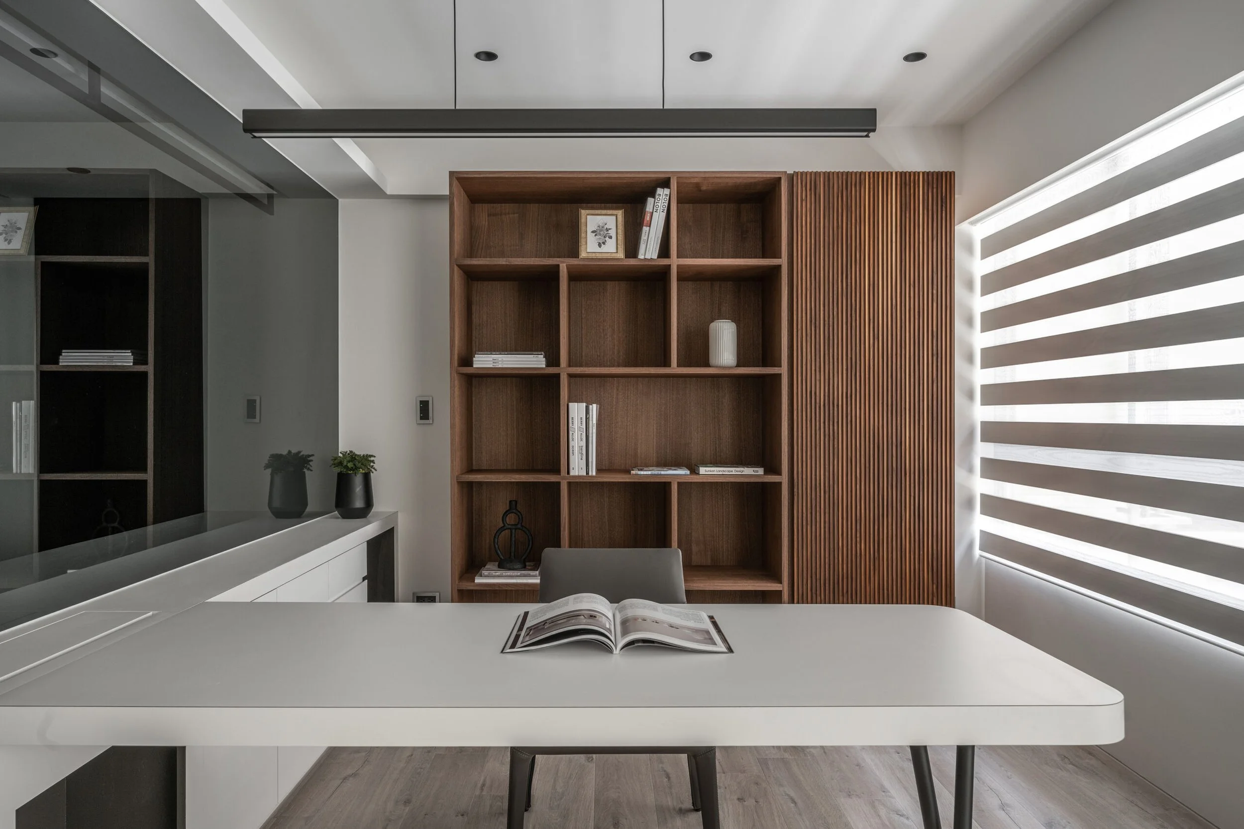 Modern dining room with a white table, open magazine, wooden bookshelf, and view of wooden slatted wall and window with striped blinds.