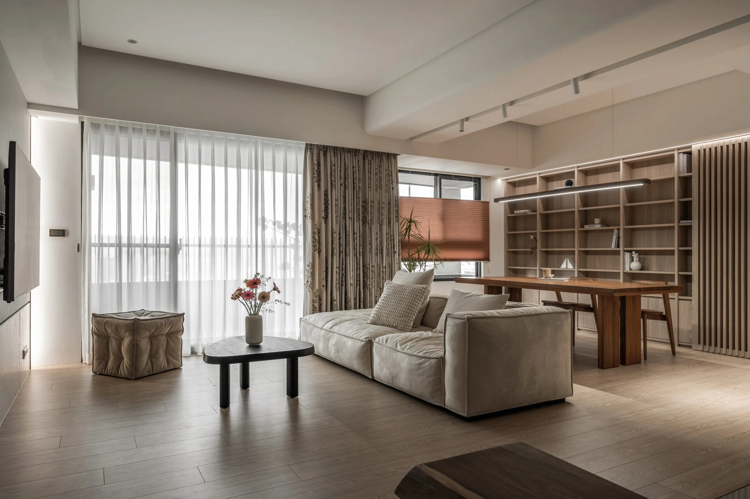 Modern living room with beige sofa, armchair, wooden coffee table, large windows with sheer white curtains, bookshelves, indoor plants, and hardwood flooring.