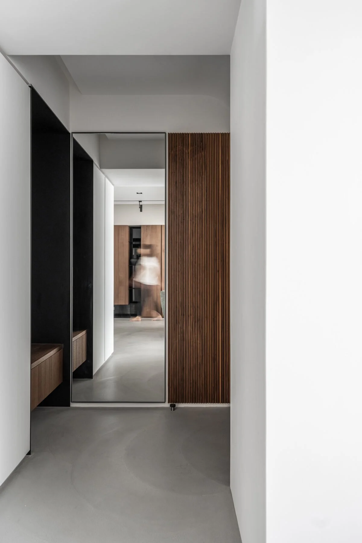 Minimalist hallway with a large mirror, white walls, wooden accents, and a blurred figure reflected in the mirror.