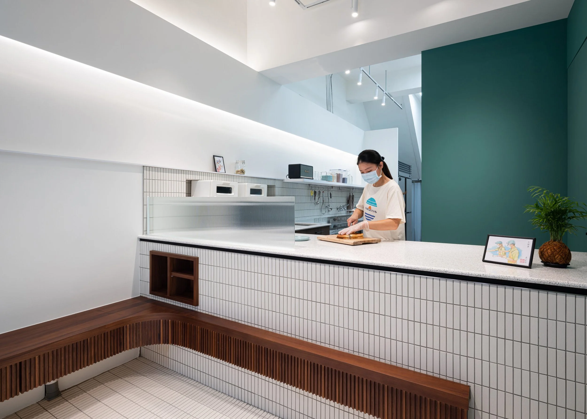 A woman with dark hair, wearing a face mask and a t-shirt, is chopping food on a wooden cutting board in a modern, bright kitchen with white tiled walls, a green accent wall, and minimalistic decor.