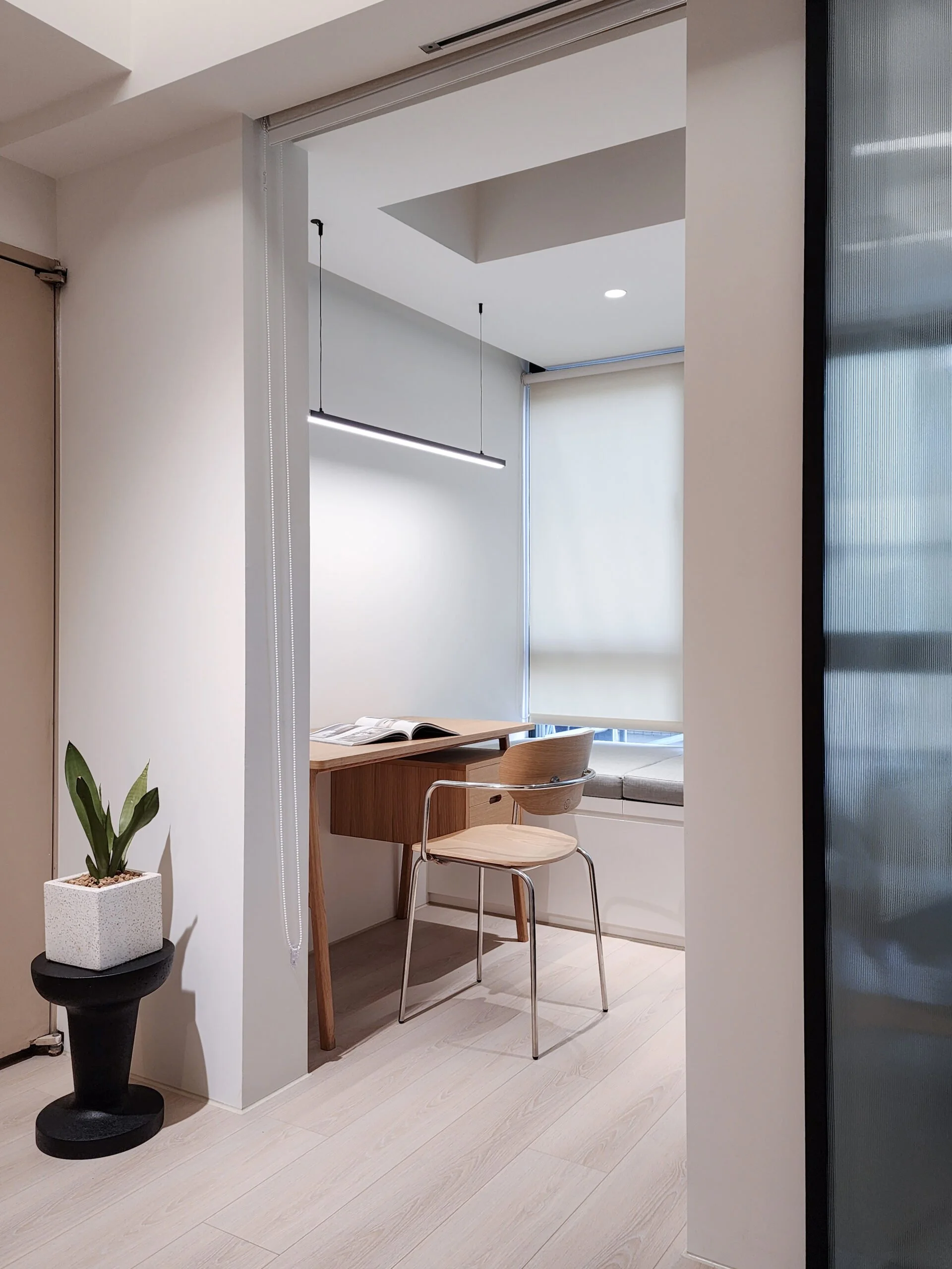 A modern interior room with minimalist decor, a small wooden table and a beige chair, a potted plant on a black stand, and a large window with a light roller blind.
