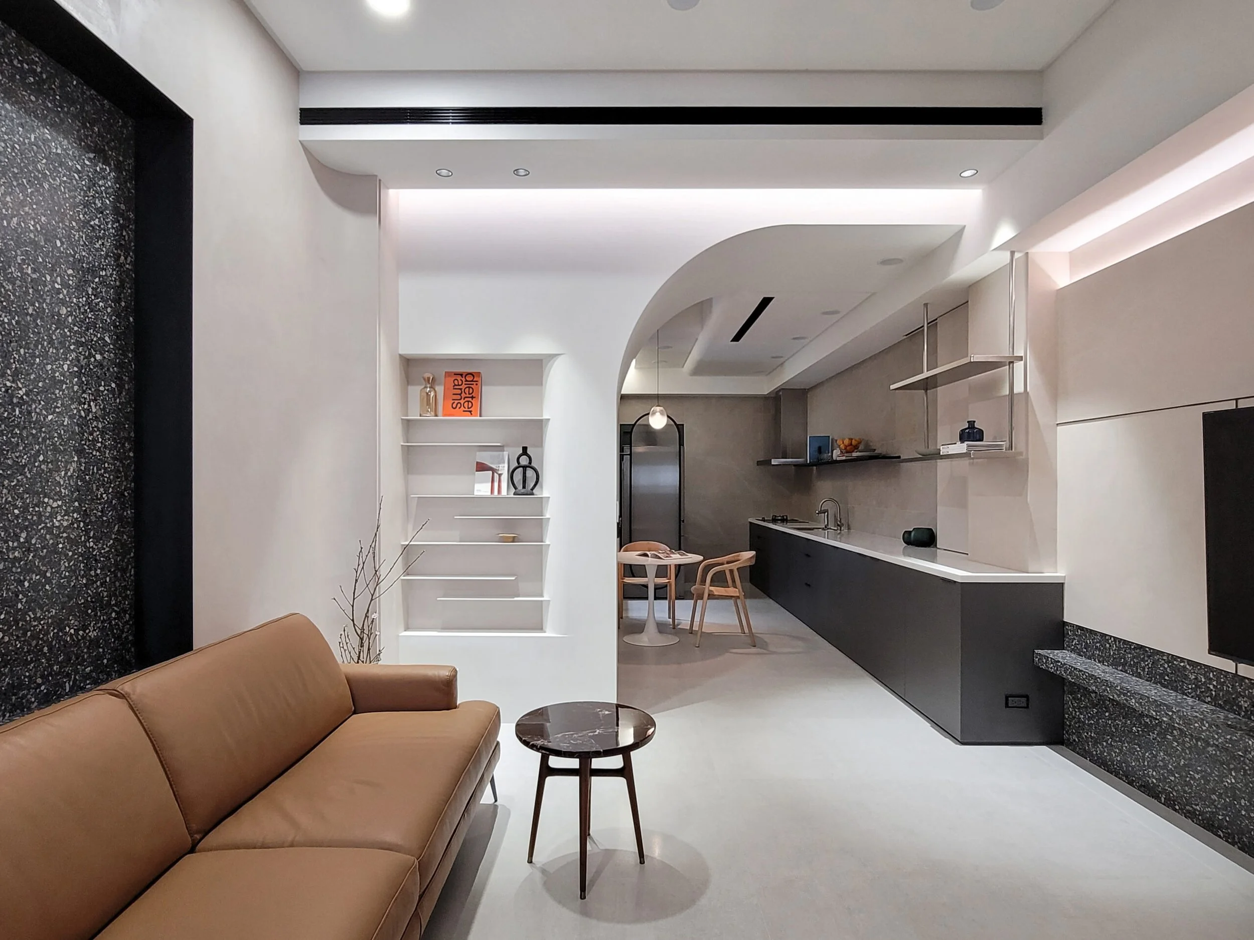 Modern living space with a brown leather sofa, a small round marble-top table with black legs, and open kitchen area with white and black cabinetry, shelves, pendant light, and decorative items.