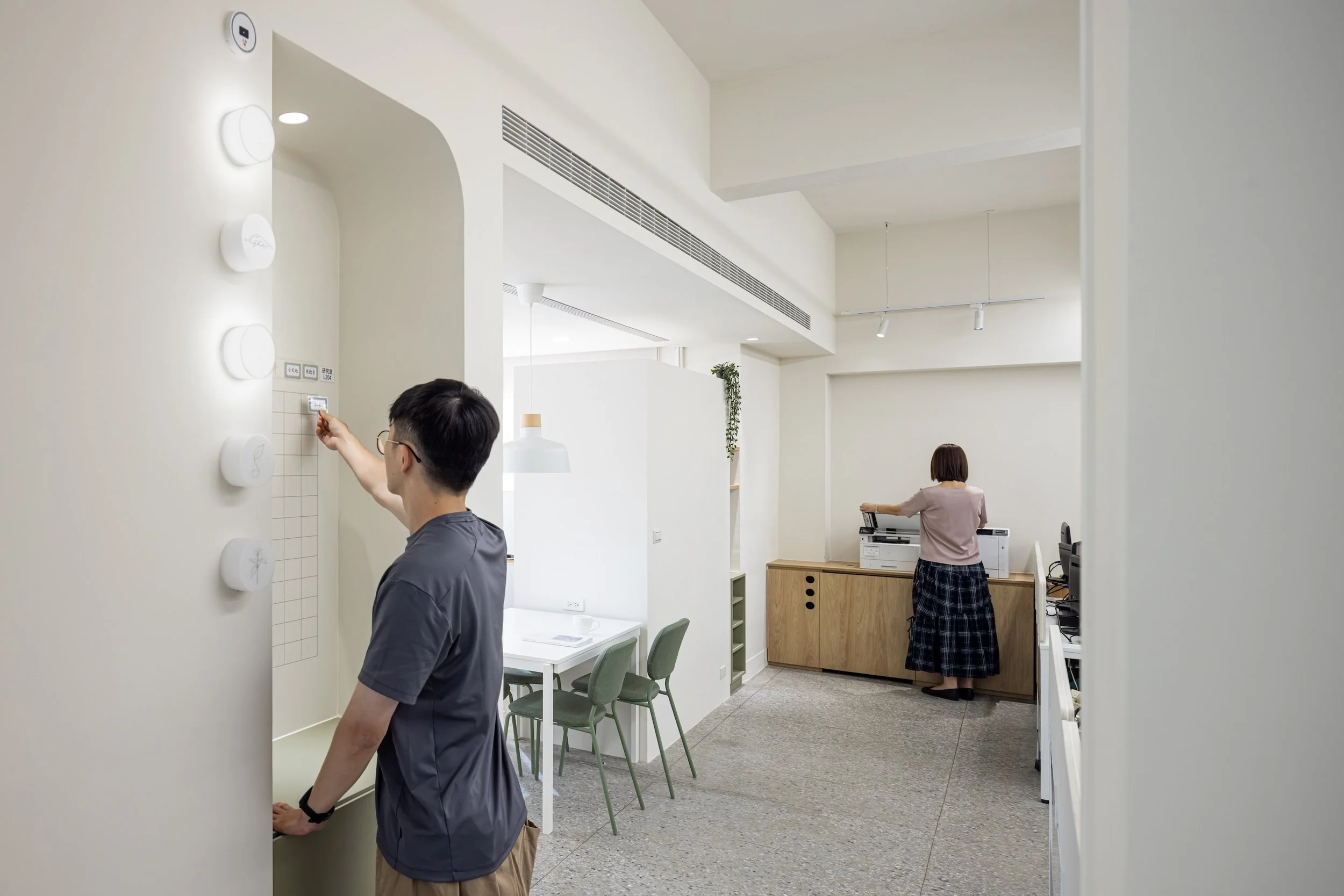 Two people are in a modern, minimalistic office or reception area. One person, with short dark hair and glasses, is standing near a wall with decorative white discs and a small grid of papers, touching or pointing to it. The other person, with a bob haircut, is wearing an apron and standing at a desk with office equipment. The space has white walls, a gray carpeted floor, green chairs, a wooden sideboard, and a hanging white lamp. There are indoor plants and track lighting.