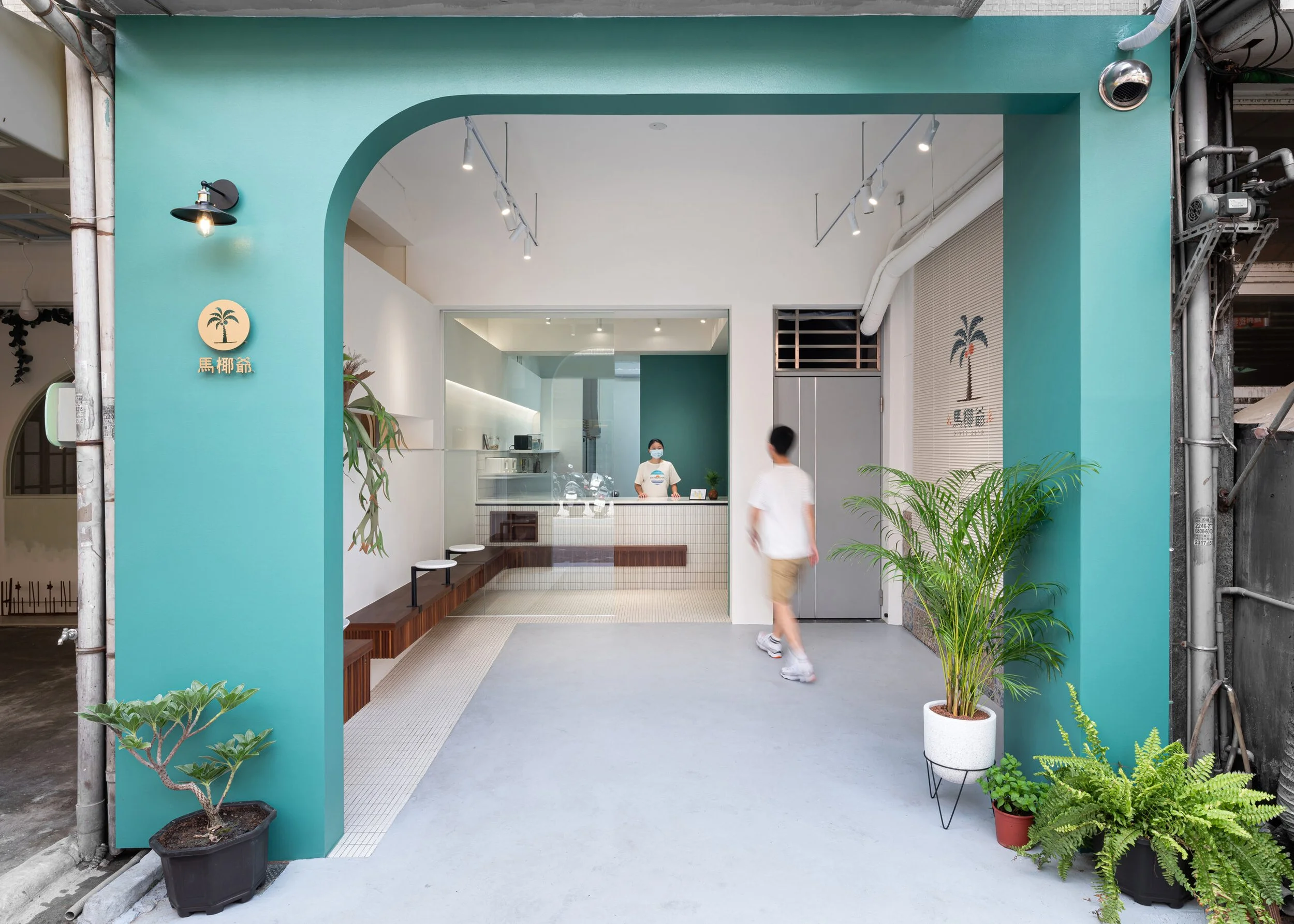 Modern storefront with turquoise walls and an arched entrance, potted plants outside, and two people inside, one wearing a mask, in a bright, minimal interior.