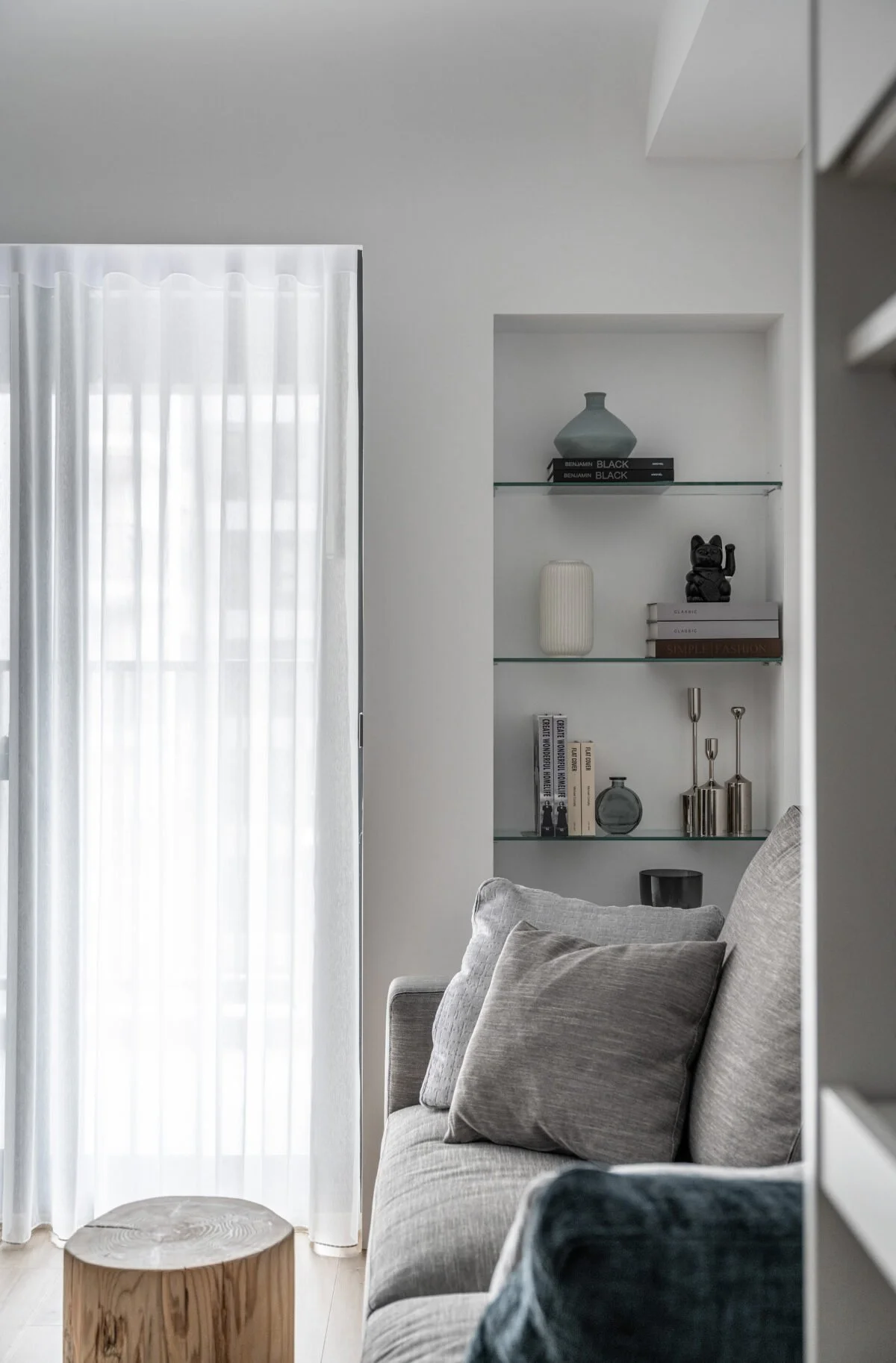 Corner of a living room with a light gray sofa and two pillows, a small wooden stool in front, a built-in wall shelf with decorative items and books, and a window with white curtains