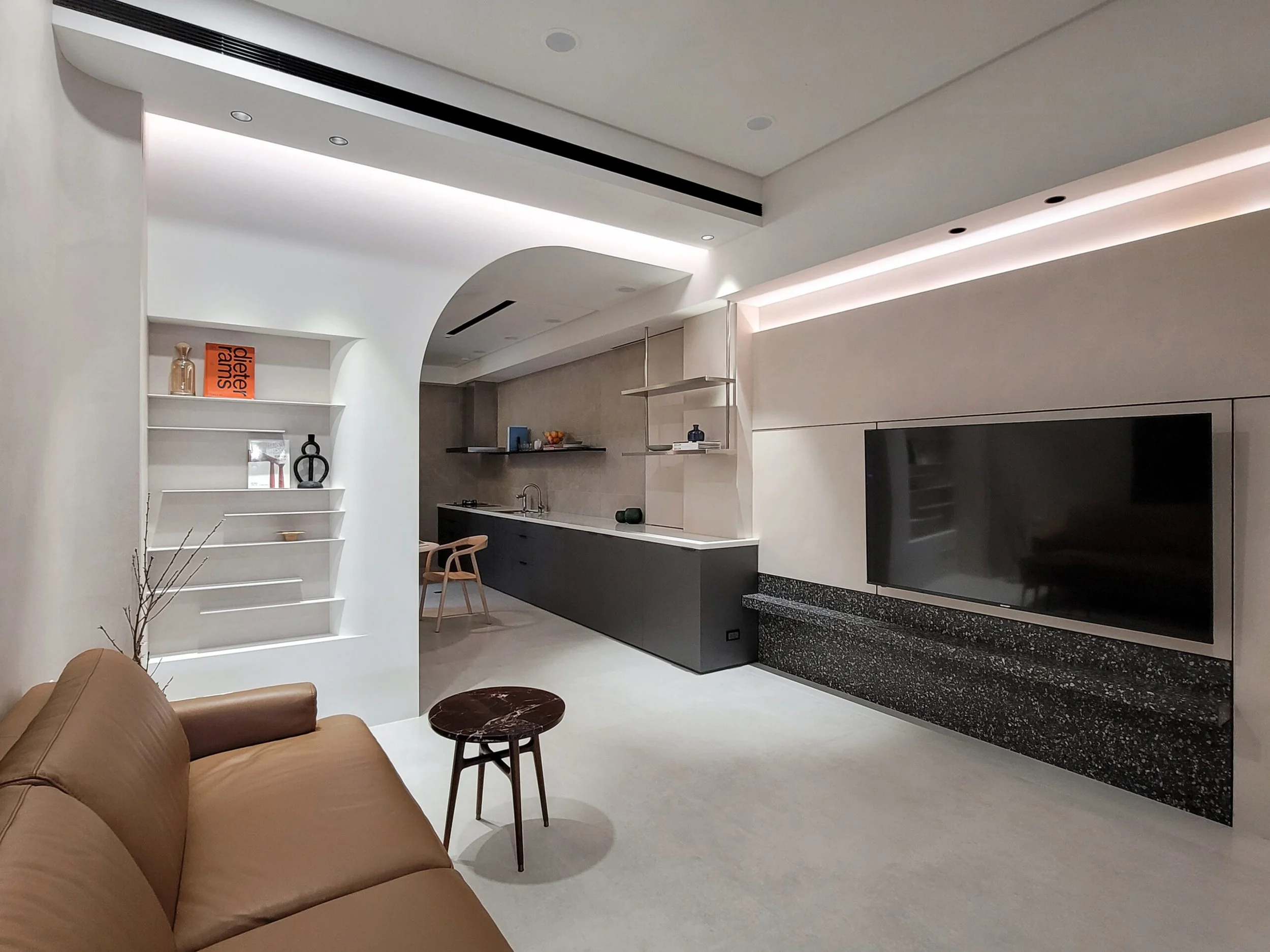 Modern living room with a tan leather sofa, small side table, white walls, open kitchen in the background with floating shelves, black cabinetry, and a flat-screen TV.