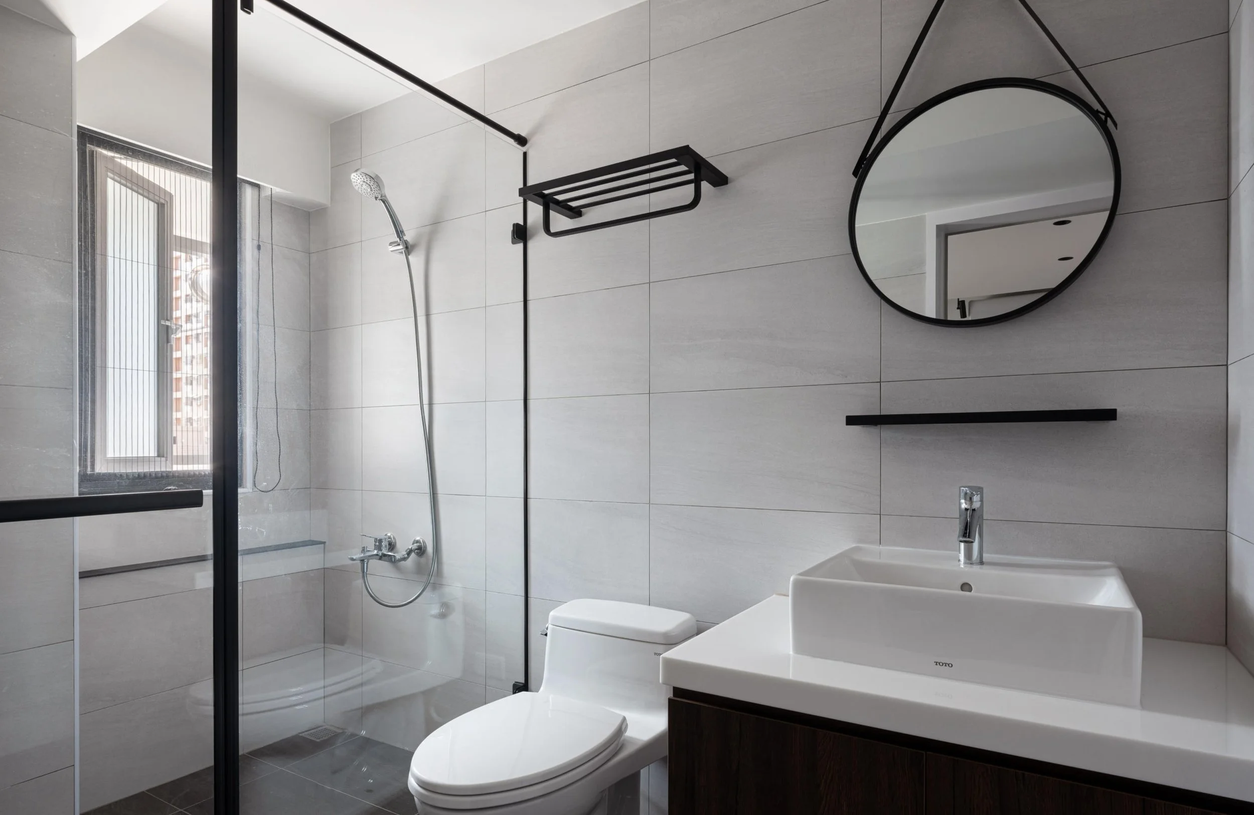 Modern bathroom with gray tiled walls, glass shower enclosure, wall-mounted shower head, round mirror, white sink, and a toilet.