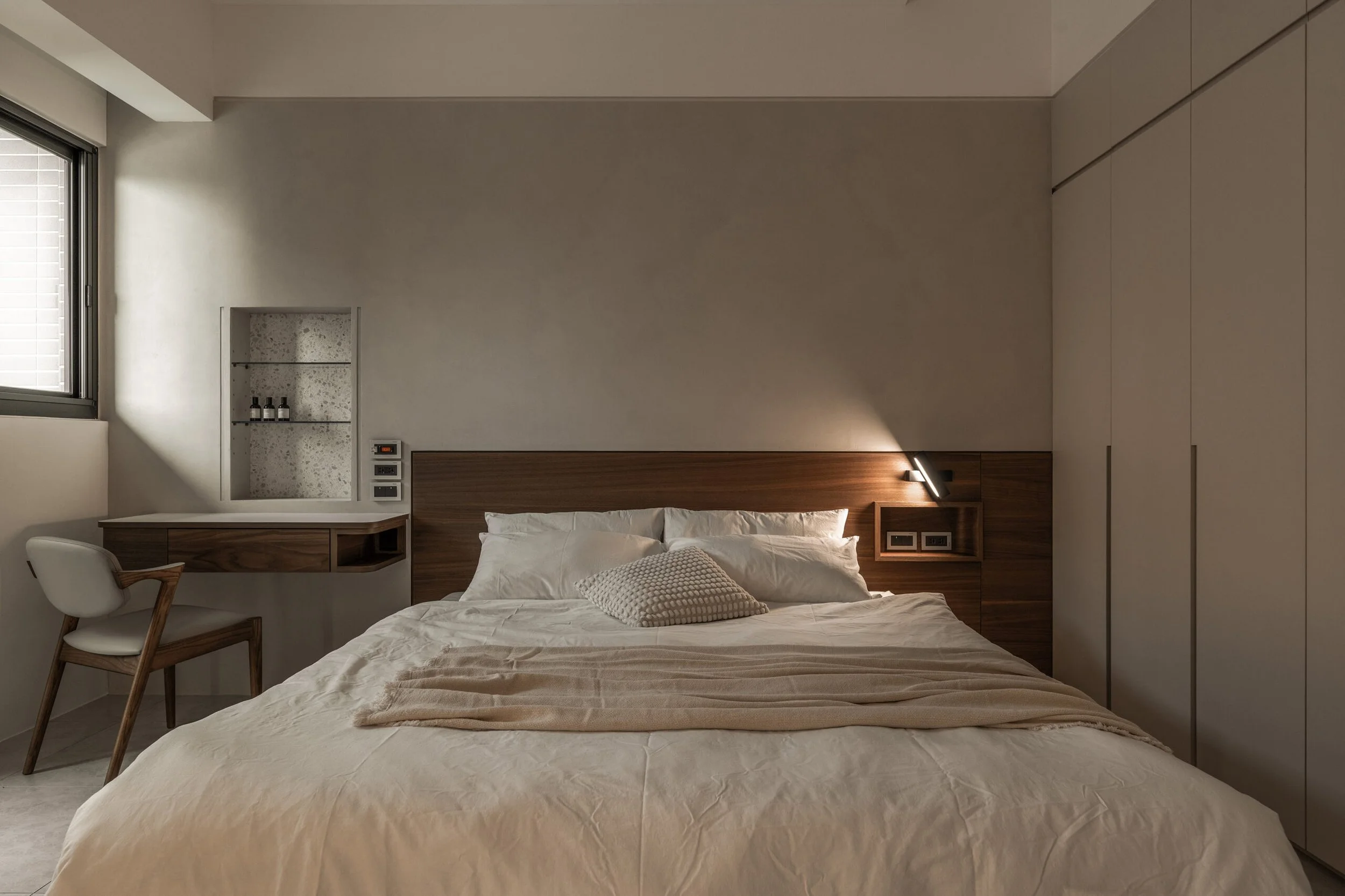 Modern bedroom with beige walls, a large bed with white bedding and pillows, a wooden headboard with built-in nightstands and outlets, and a textured beige blanket. To the left, a white chair with wooden arms and a small desk with shelves holding bottles. There is a window with a blind on the left wall, and built-in closets on the right.