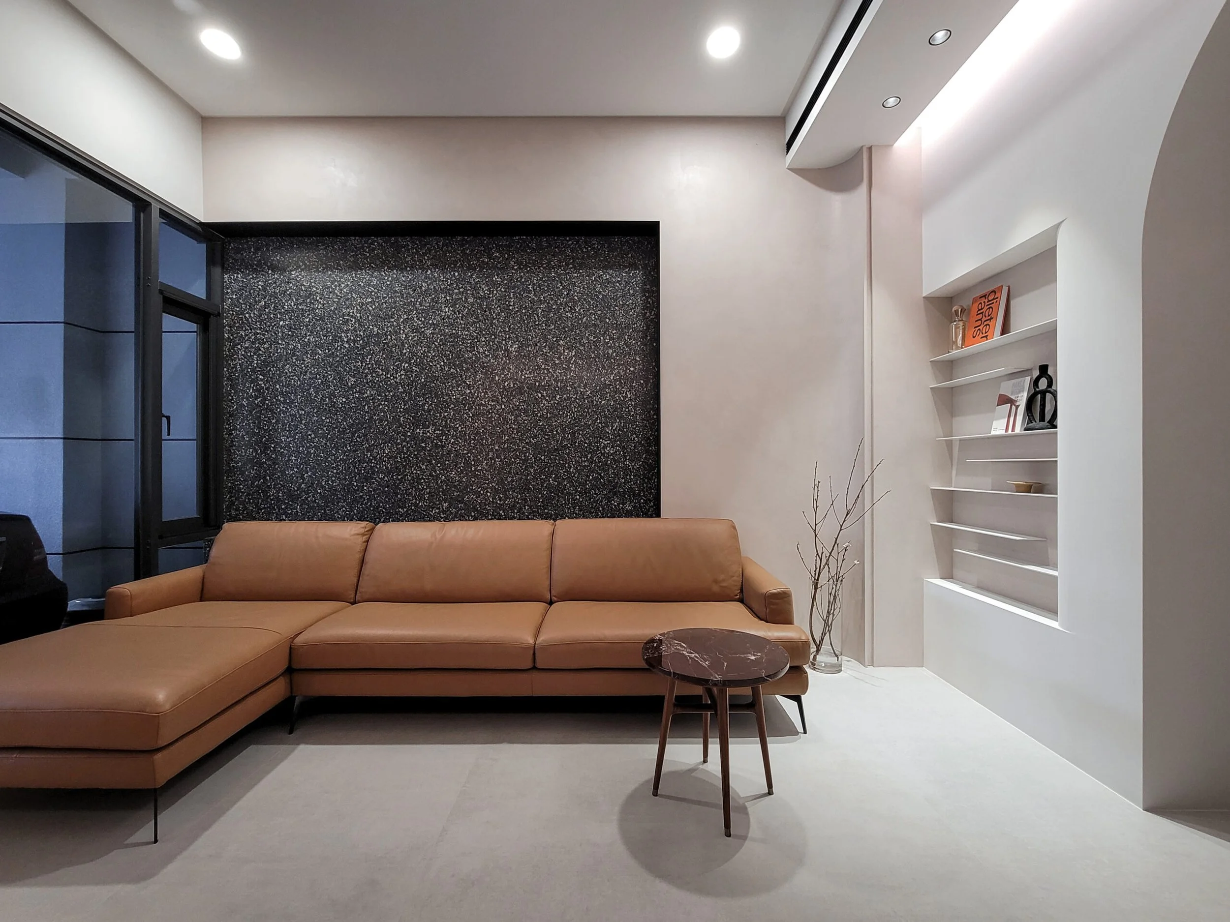 Modern interior living room with a tan leather sectional sofa, a small round side table, a white bookshelf with decorative items and books, light-colored walls, and ceiling spotlights.
