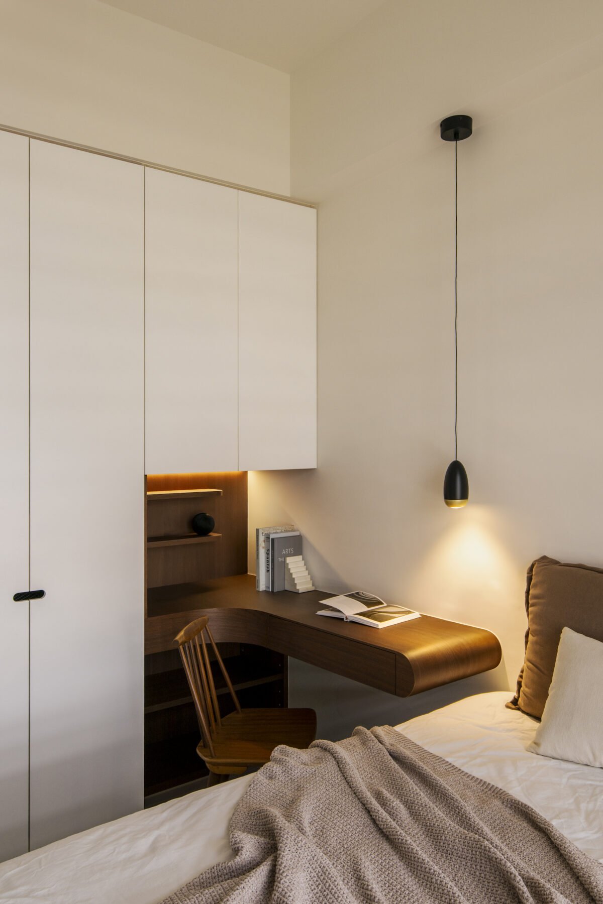 A cozy bedroom corner with a bed, a wooden desk with books, a wooden chair, wall-mounted white cabinets, a small open shelf with decorative items, and a modern black pendant light
