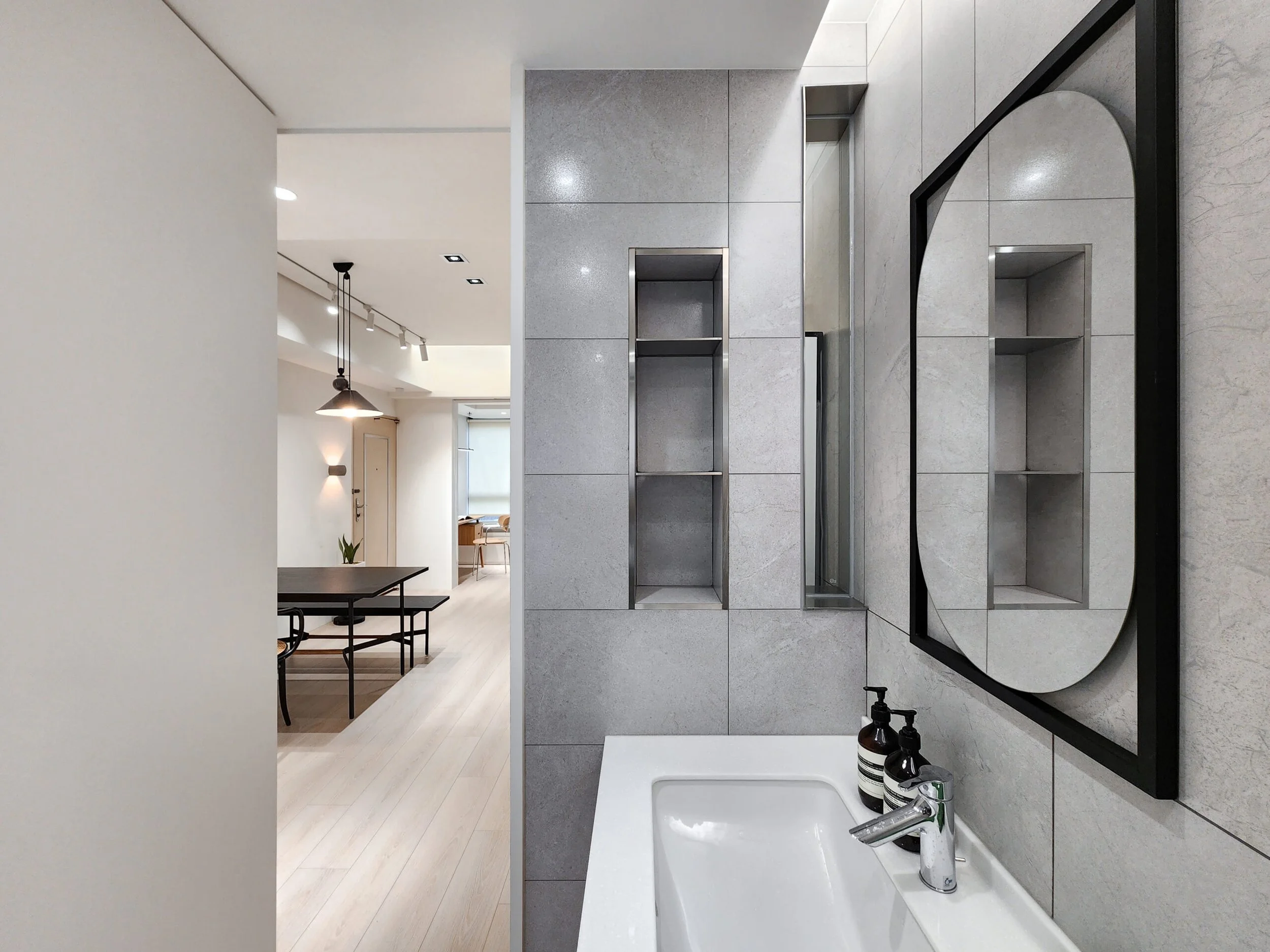 A modern bathroom with a white sink, black framed mirror, gray tiled wall, and black soap dispensers. The bathroom has light wood flooring, and in the background, there is a dining area with black chairs, a table, and hanging black pendant lights.