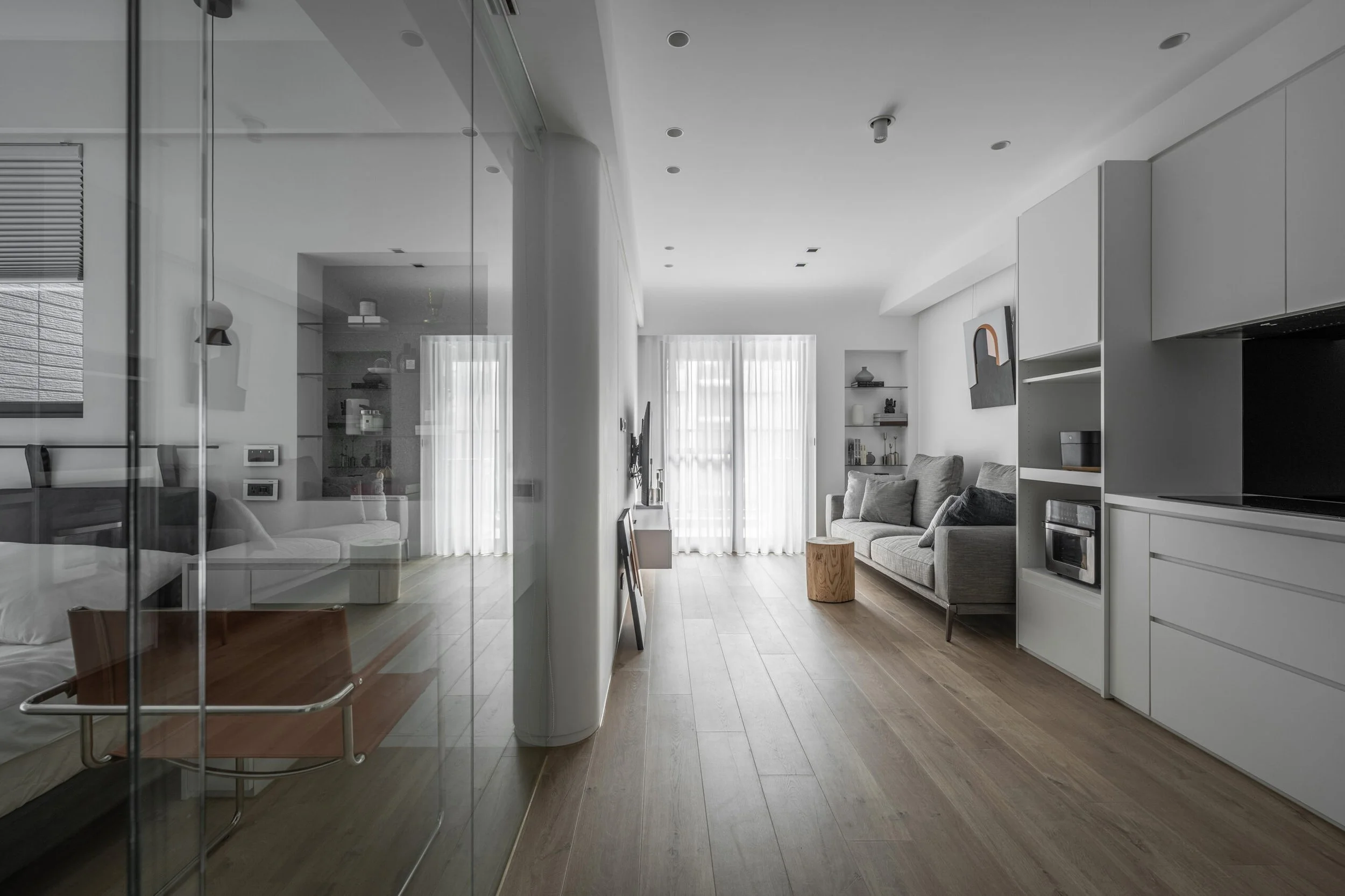Modern living room with gray sofa, wooden side table, shelves with decor, and large windows with sheer curtains, viewed through glass wall.