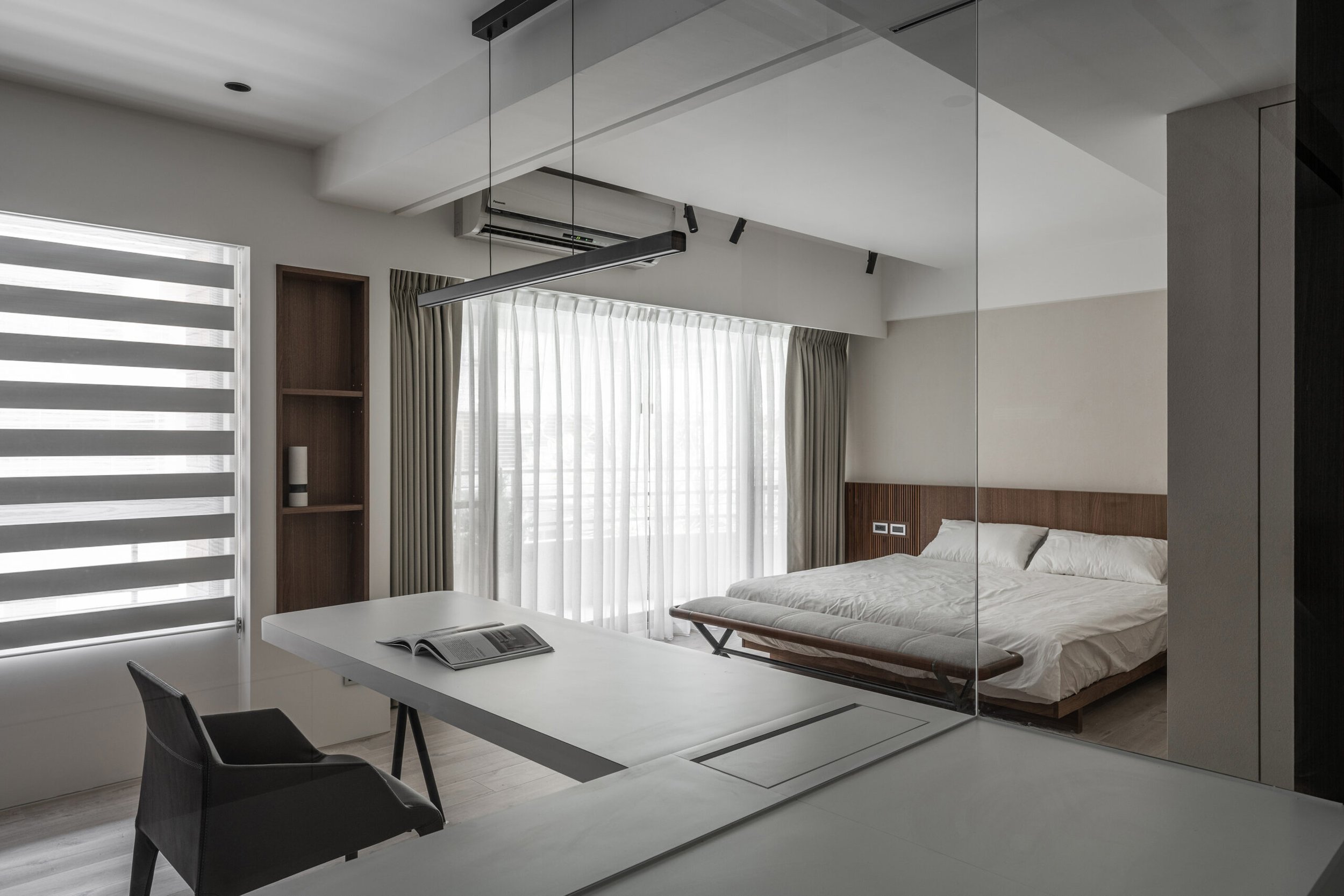 Modern bedroom with white walls, a bed with white linens, wooden headboard, and large window with sheer curtains. There is a private enclosed space visible through a glass wall with a table, book, and chairs.