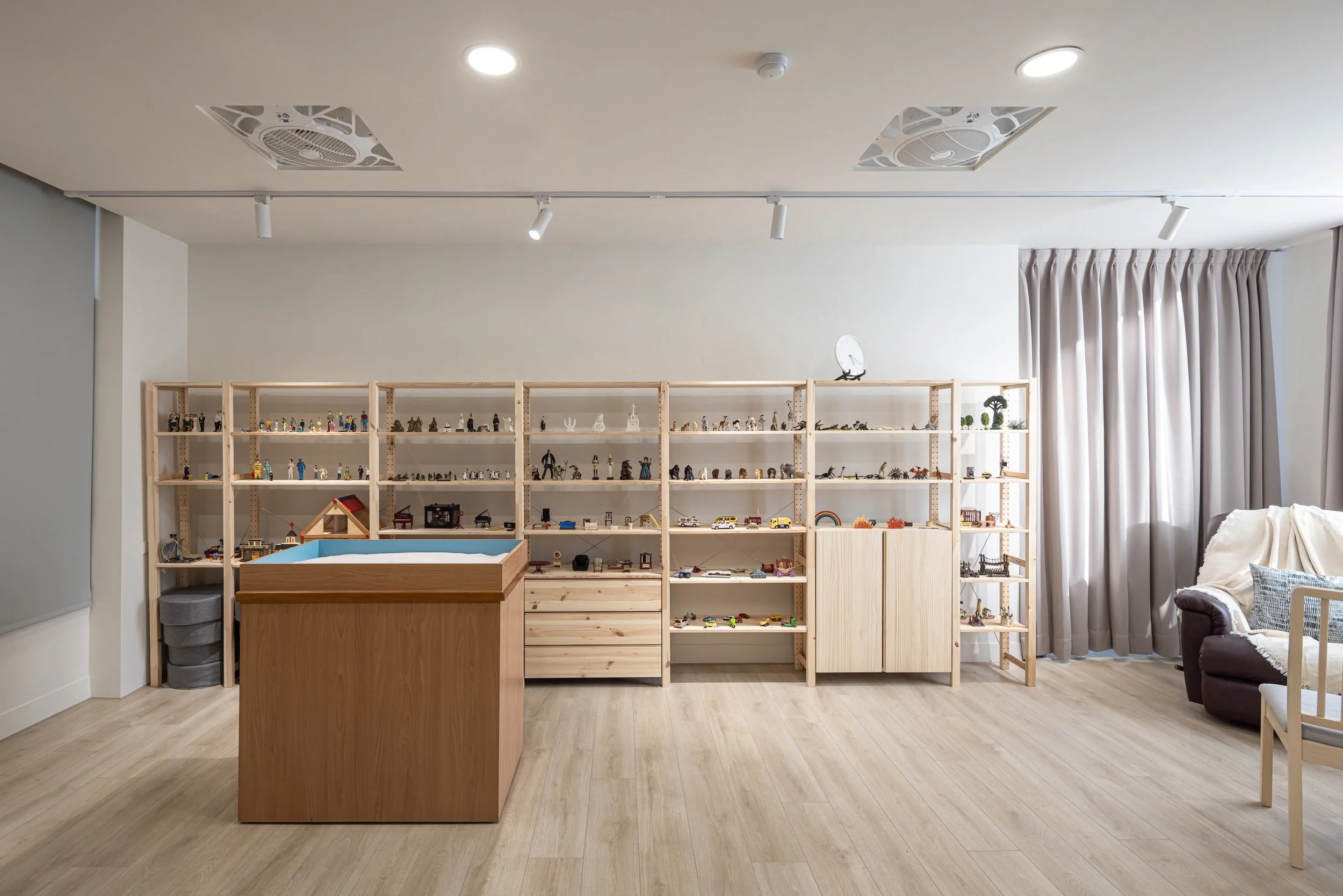 Interior of a room with wooden shelves filled with small toys and figurines, beige curtains on the right, and a small wooden table in front.