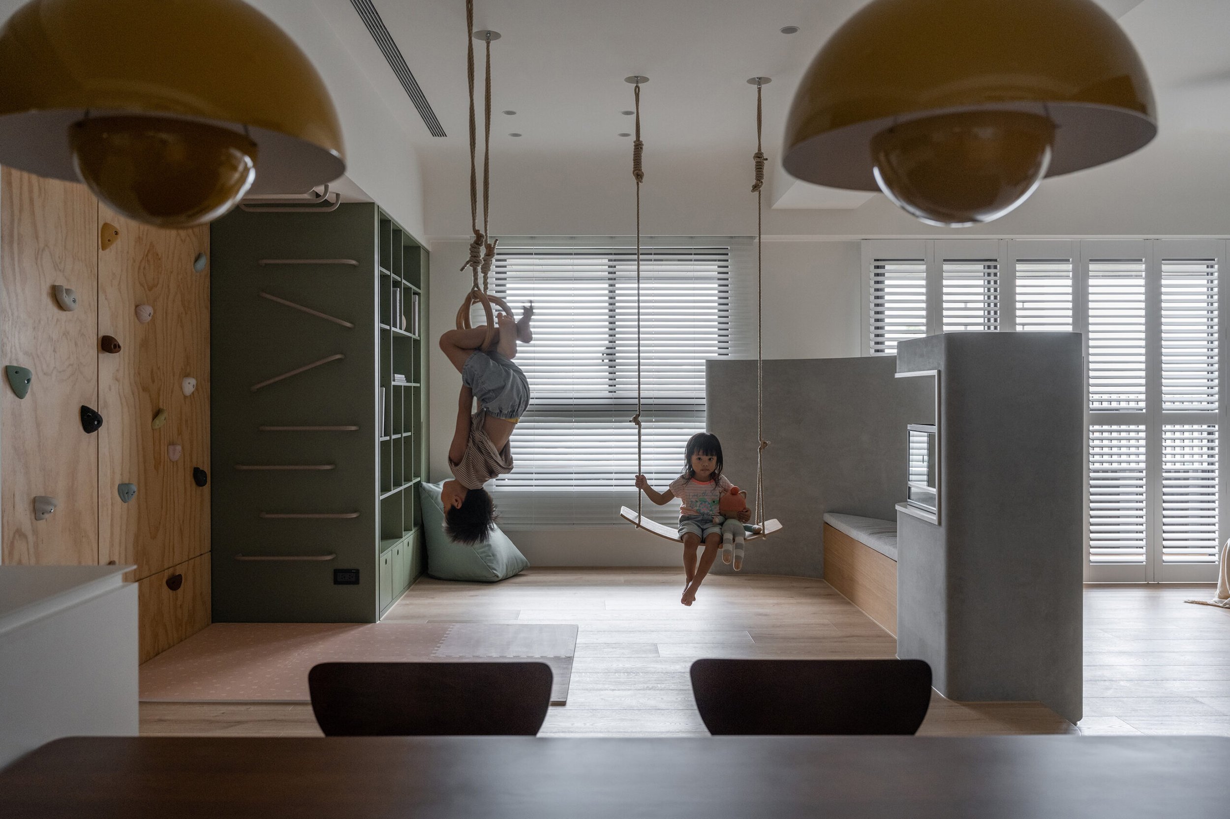 Two children playing indoors, one is hanging upside down from a rope, the other sitting on a swing, in a room with large windows, wooden floors, and modern furniture.