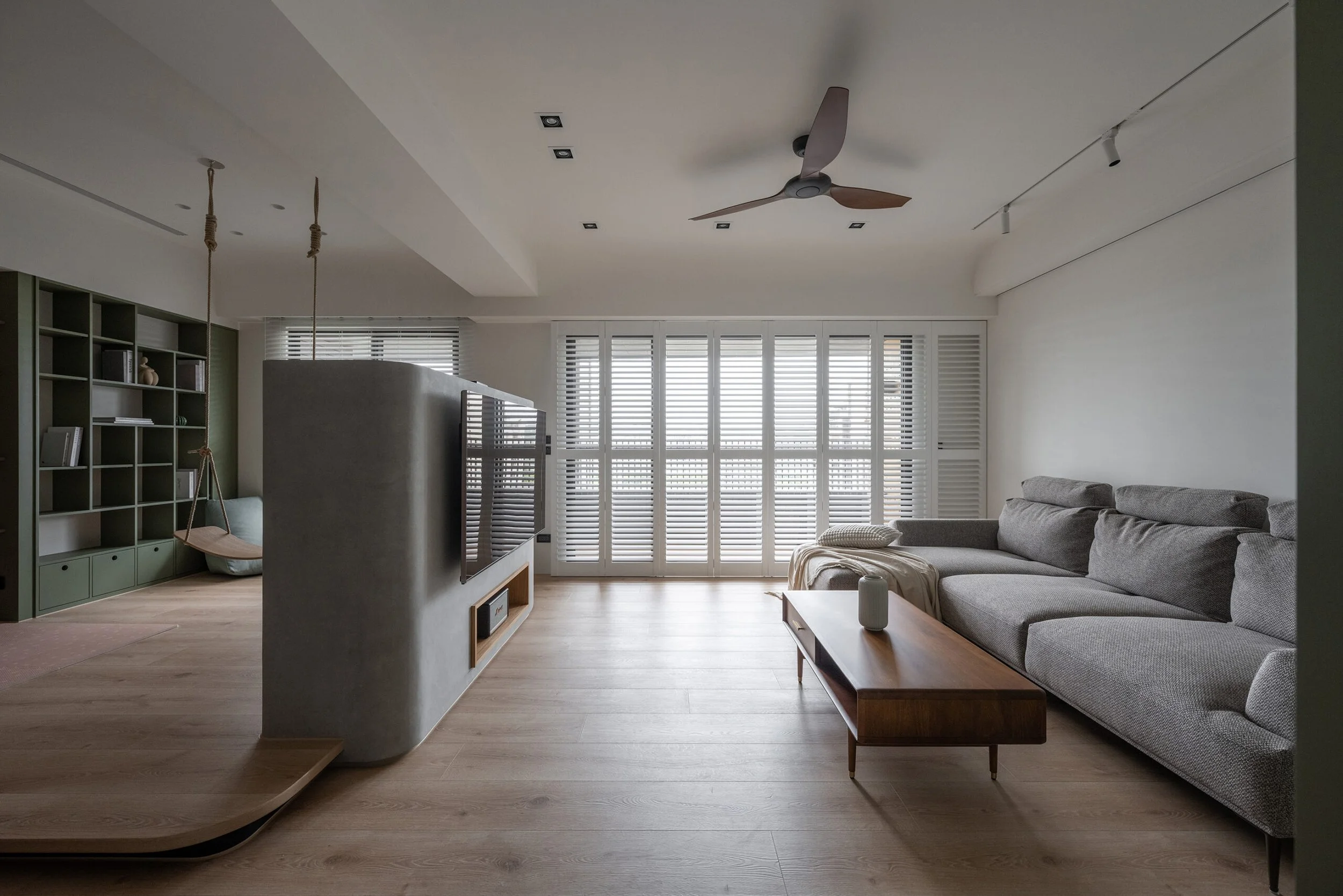Living room with a gray sofa, wooden coffee table, large window with white shutters, ceiling fan, and a built-in bookshelf with decorative items.