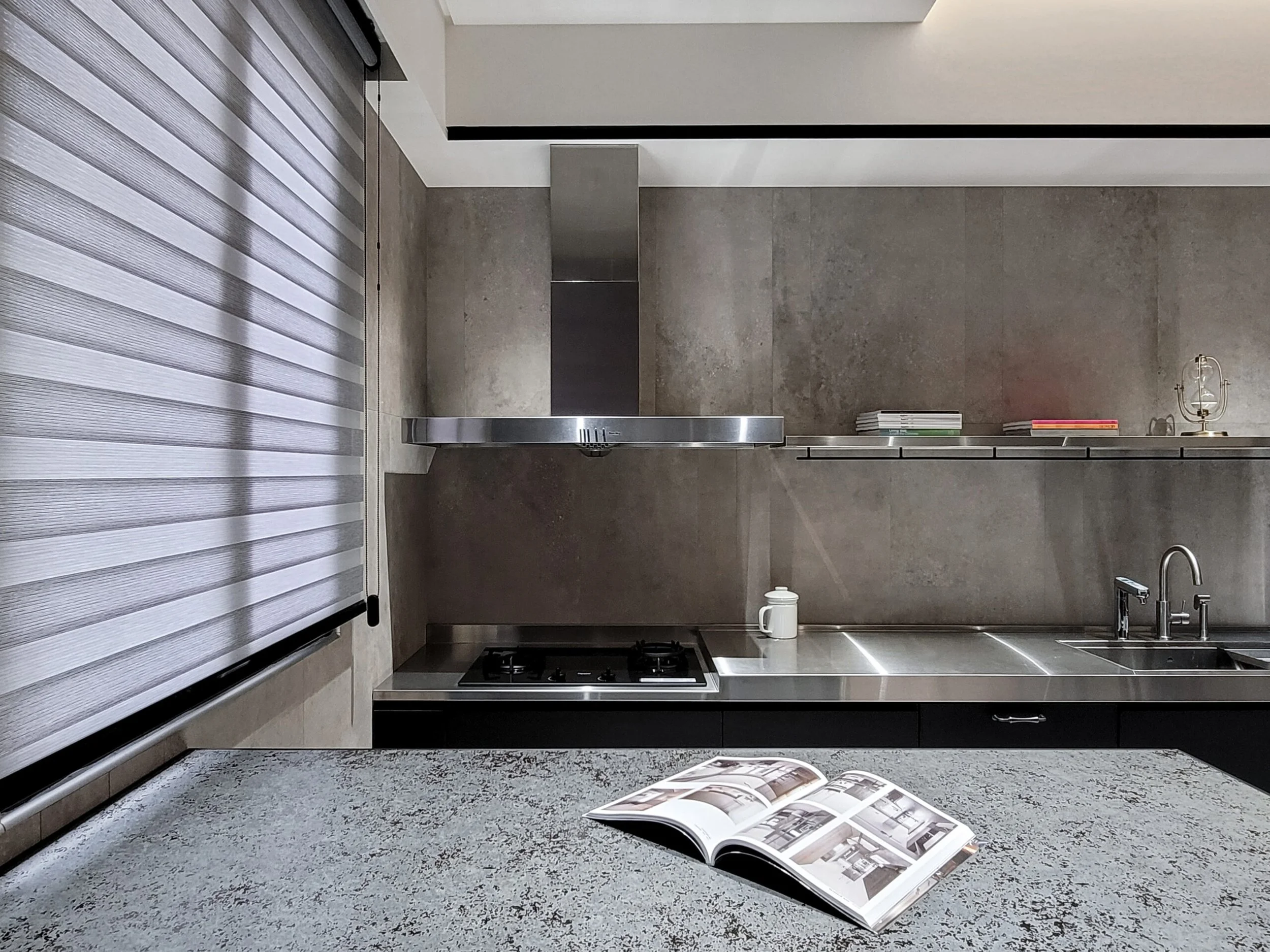 Modern kitchen with gray cabinets, a stainless steel range hood, a gas stove, and an open book on a speckled gray stone countertop. Books are on a shelf above the counter, and a window with a striped roller blind is on the left.