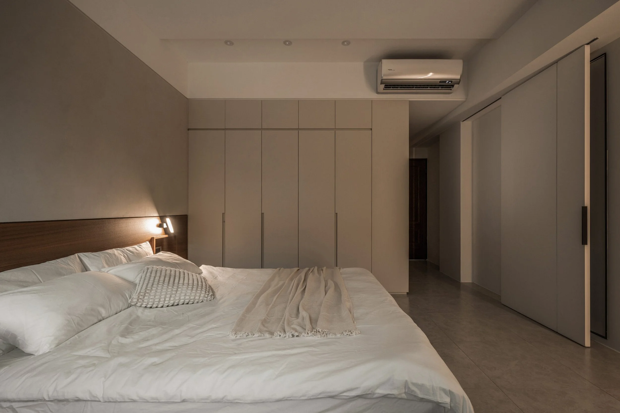 A modern bedroom with a bed, white bedding, and decorative pillows, large built-in wardrobe, wall-mounted light, and an air conditioning unit.