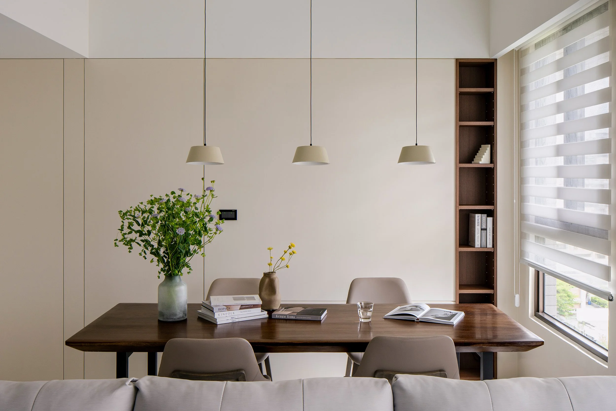 Modern dining room with a wooden table, three beige chairs, a glass of water, books, and two decorative vases with flowers. A large window with striped blinds is on the right, and a built-in wooden shelving unit is on the right wall. Three pendant lights hang above the table.