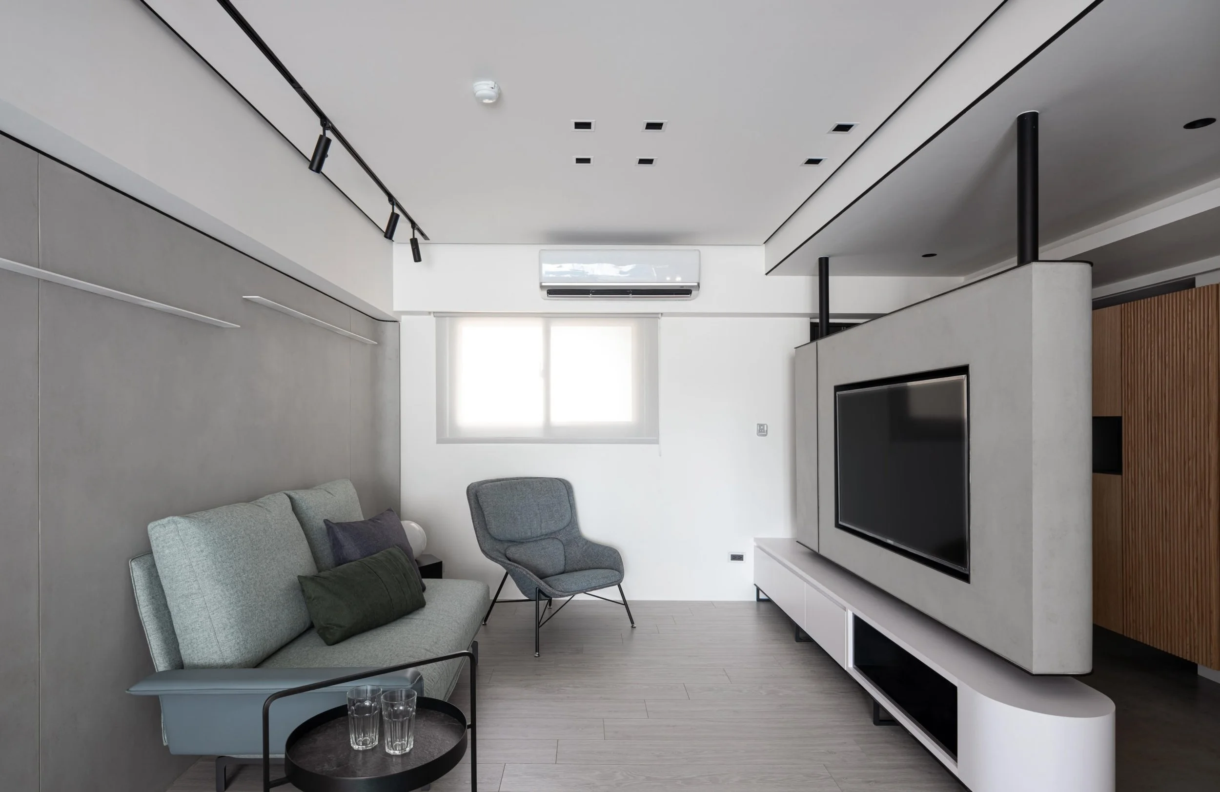 Modern living room with a light gray sofa, two green pillows, and a gray armchair, a black round side table with two glasses, a large flat-screen TV on a white entertainment unit, and a ceiling white air conditioner, window with lights, and track lighting.