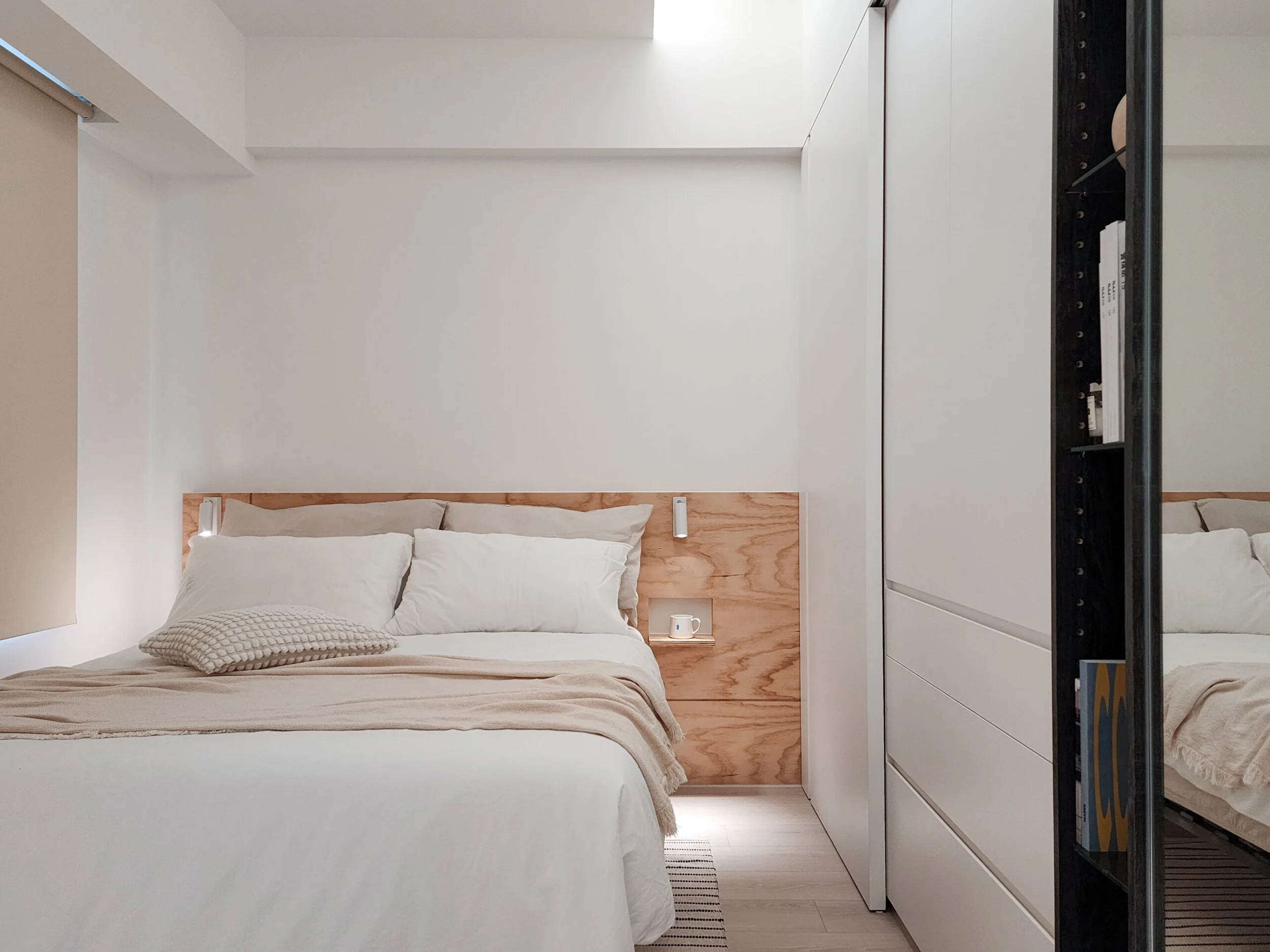 A minimalistic bedroom with a white bed, beige blanket, and a textured beige pillow. The headboard is made of light wood, and there's a small white nightstand with a white mug and a small light attached to the wall. The wall behind the bed is plain white. To the right, white built-in drawers and a black shelving unit are visible.