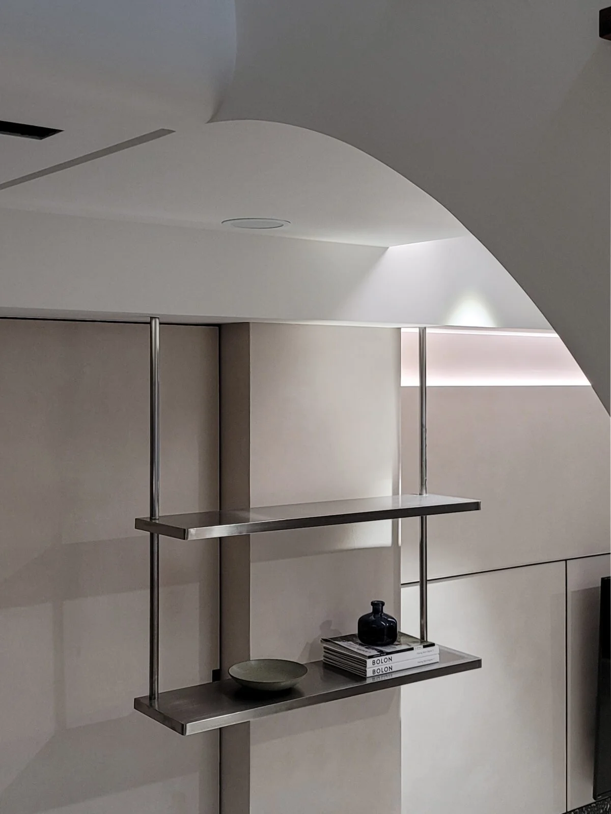 Modern minimalist wall shelf with metal frame, holding a black vase, a stack of magazines titled 'BOLON' and a small gray decorative object, against white wall with soft lighting.