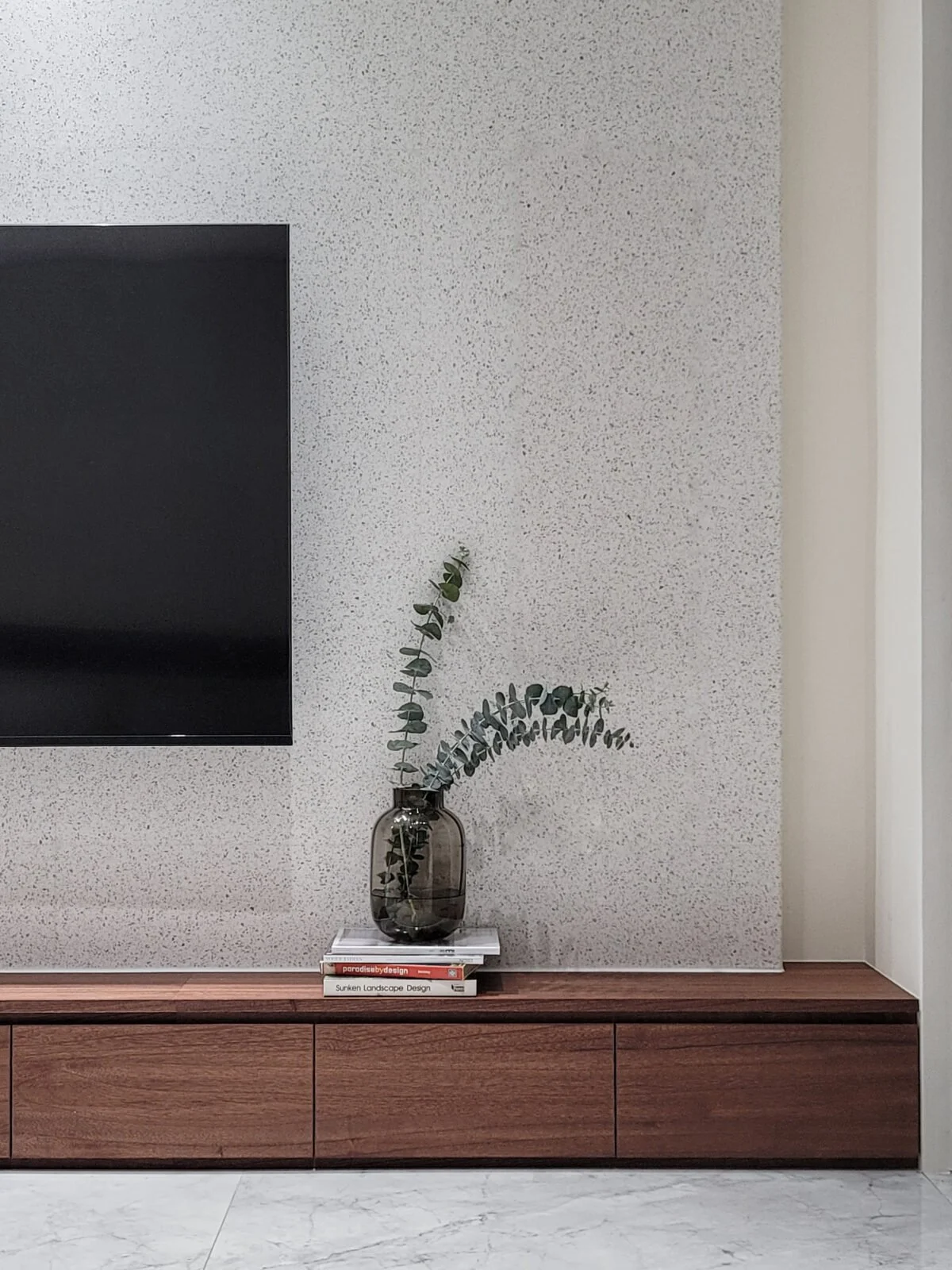 A living room wall with a flat-screen TV, a wooden console, a glass vase with eucalyptus branches, and a stack of books on top of the console.
