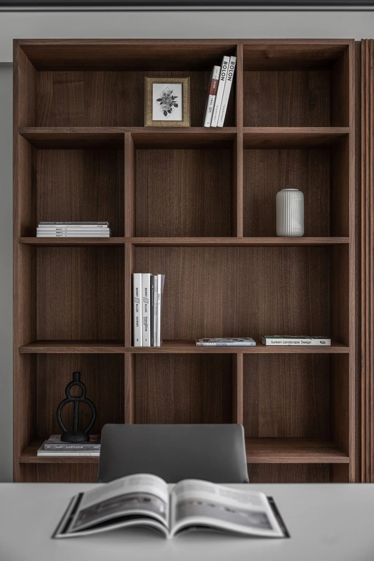 Empty wooden bookshelf with a few decorative objects, books, and a picture frame, seen from a room with a table in the foreground and an open magazine on the table.