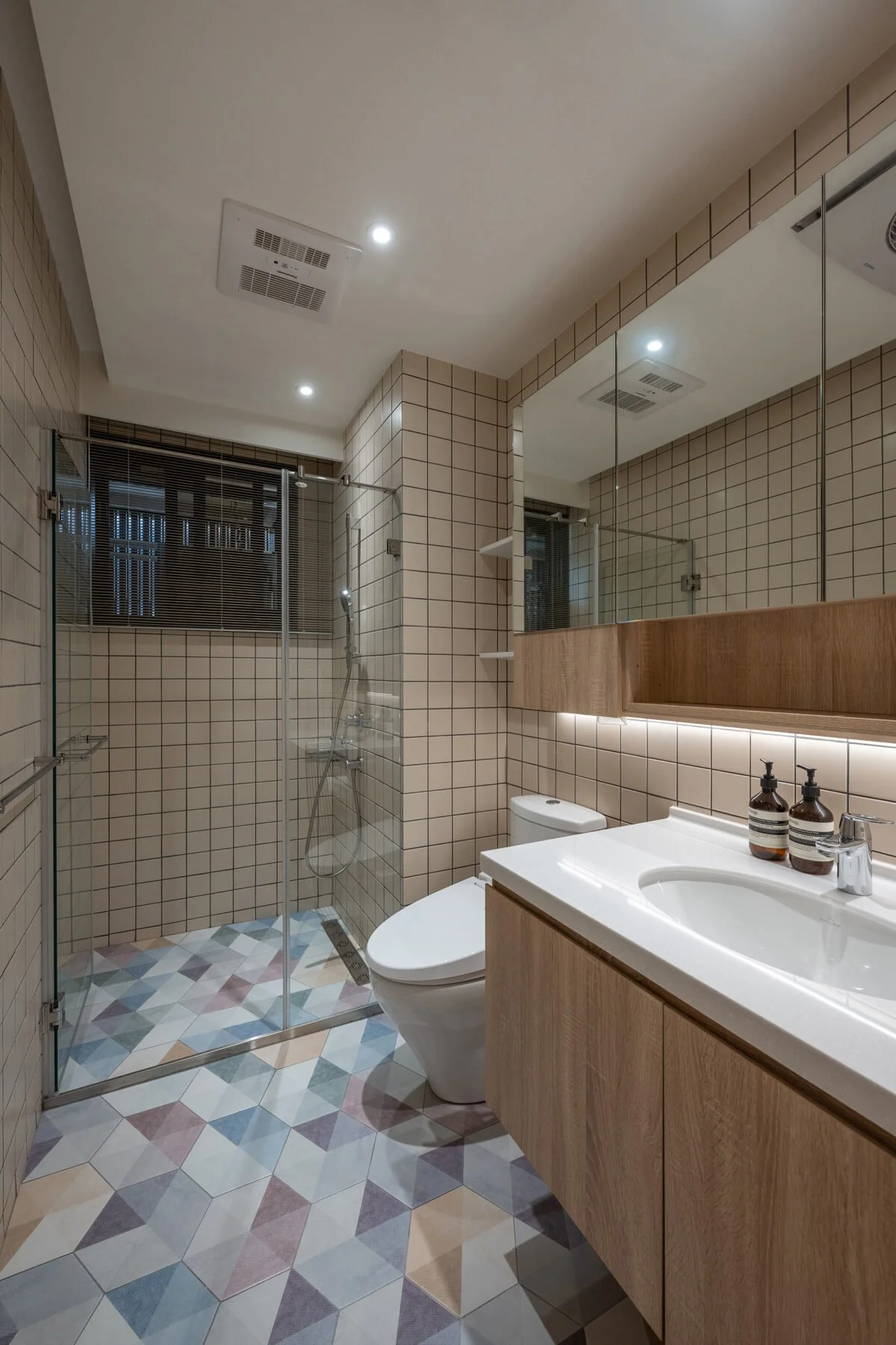 Modern bathroom with a walk-in glass shower, toilet, and wooden vanity with a white sink, two bottles of liquid soap, a large mirror, beige tiled walls, and a patterned tiled floor.